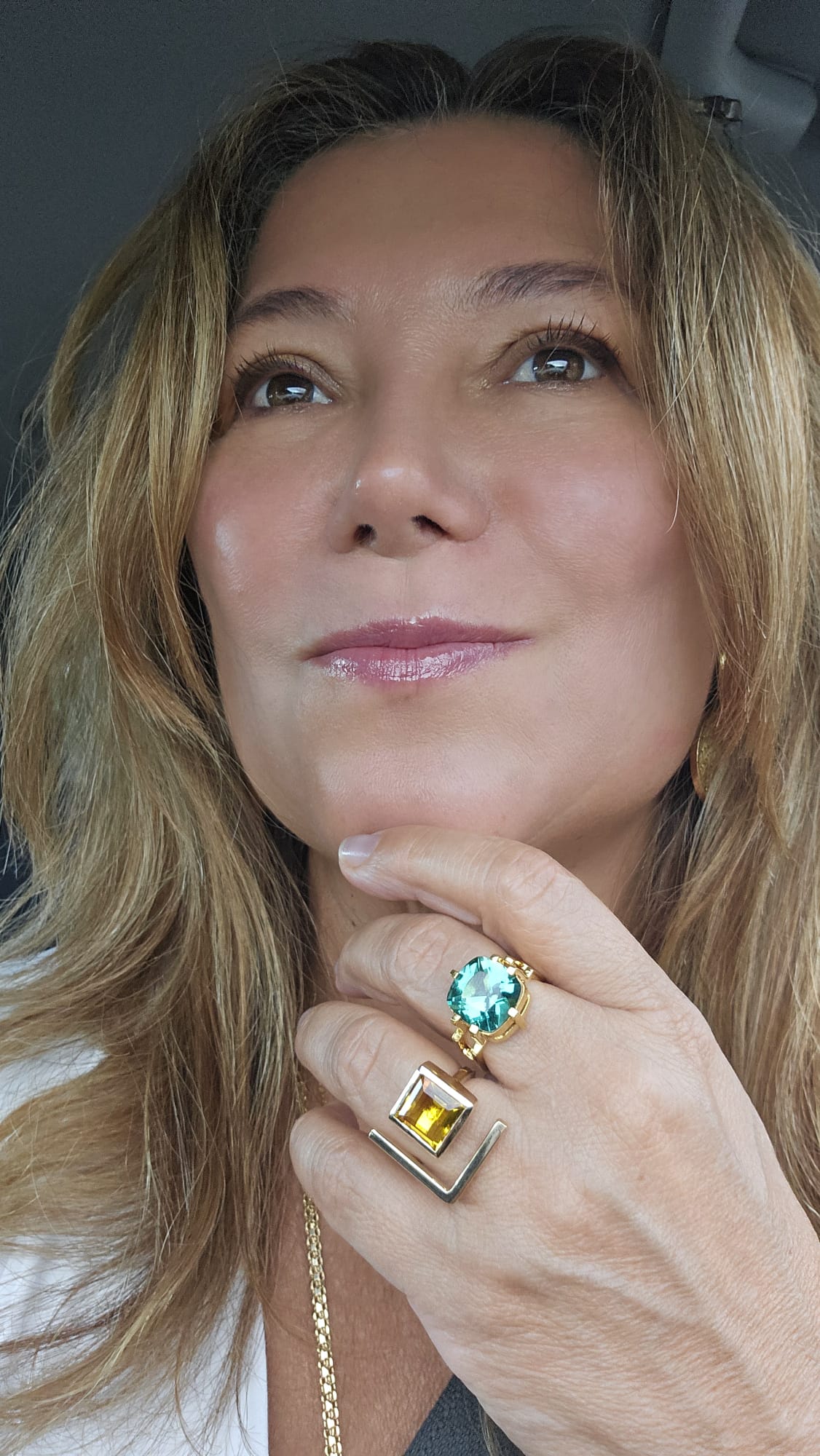 Woman with wavy hair, wearing rings and a necklace, resting her chin on her hand, looking upward.