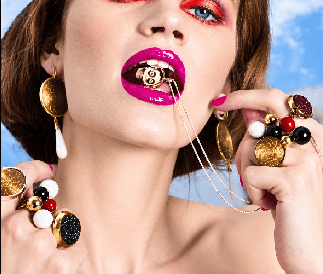 Close-up of a woman with bright makeup, gold jewelry, and colorful accessories, holding a necklace in her mouth against a blue sky background.
