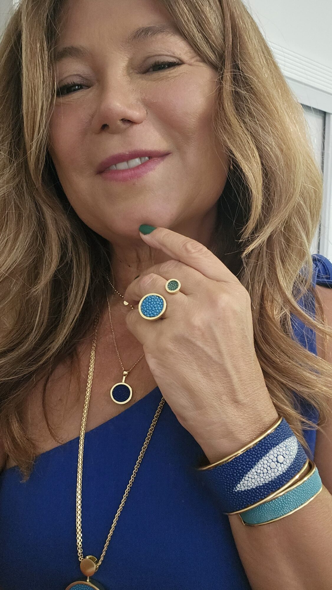 Woman with long hair wearing blue top and jewelry, smiling, touching her chin with her hand.