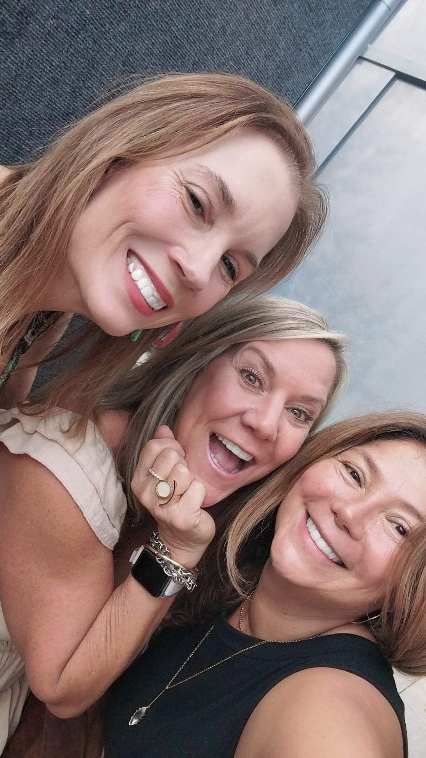Three women smiling and posing closely together outdoors, with a building in the background.