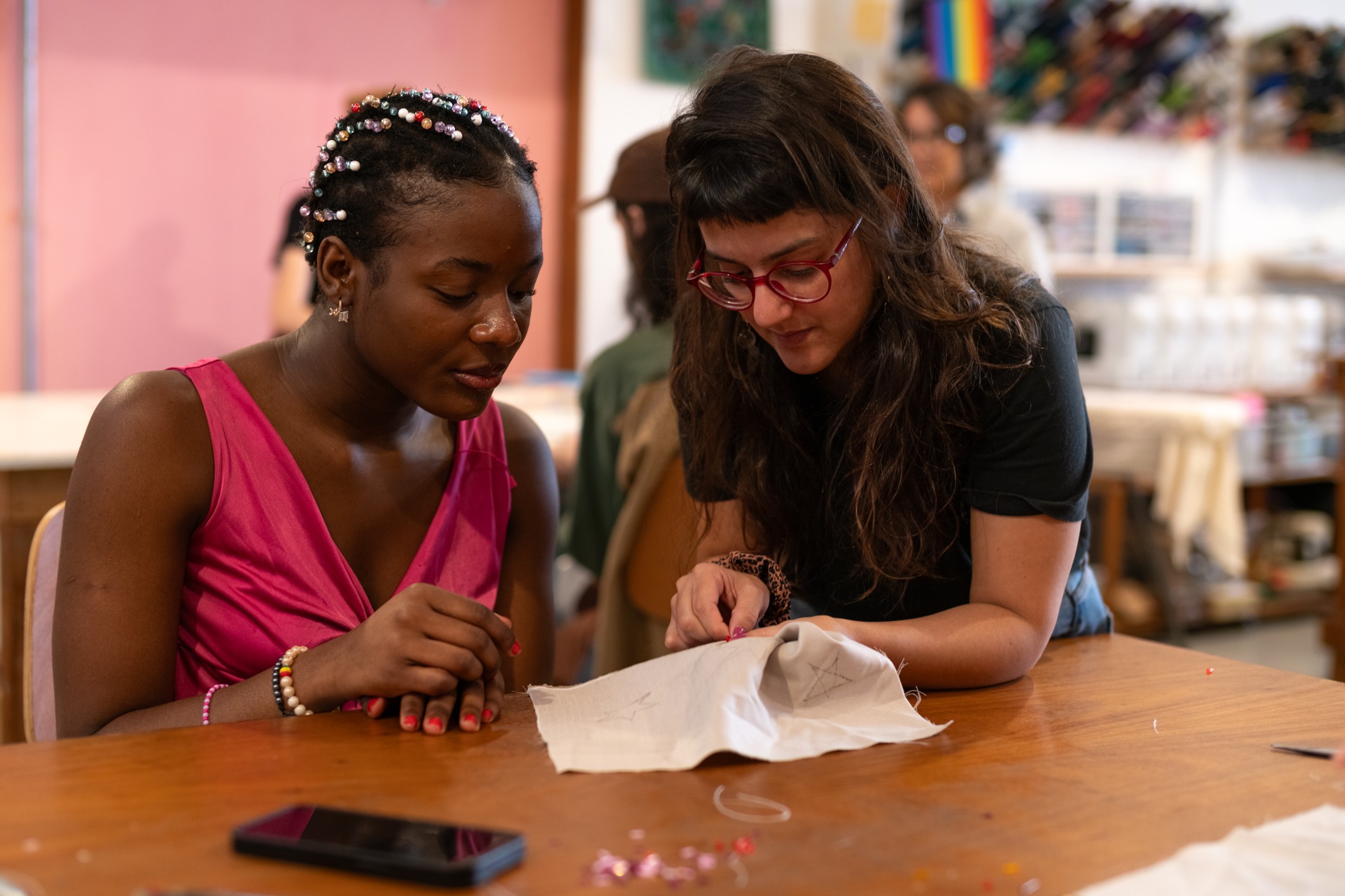 Two women sit at a table, looking at a piece of cloth or paper together in a store or craft space.