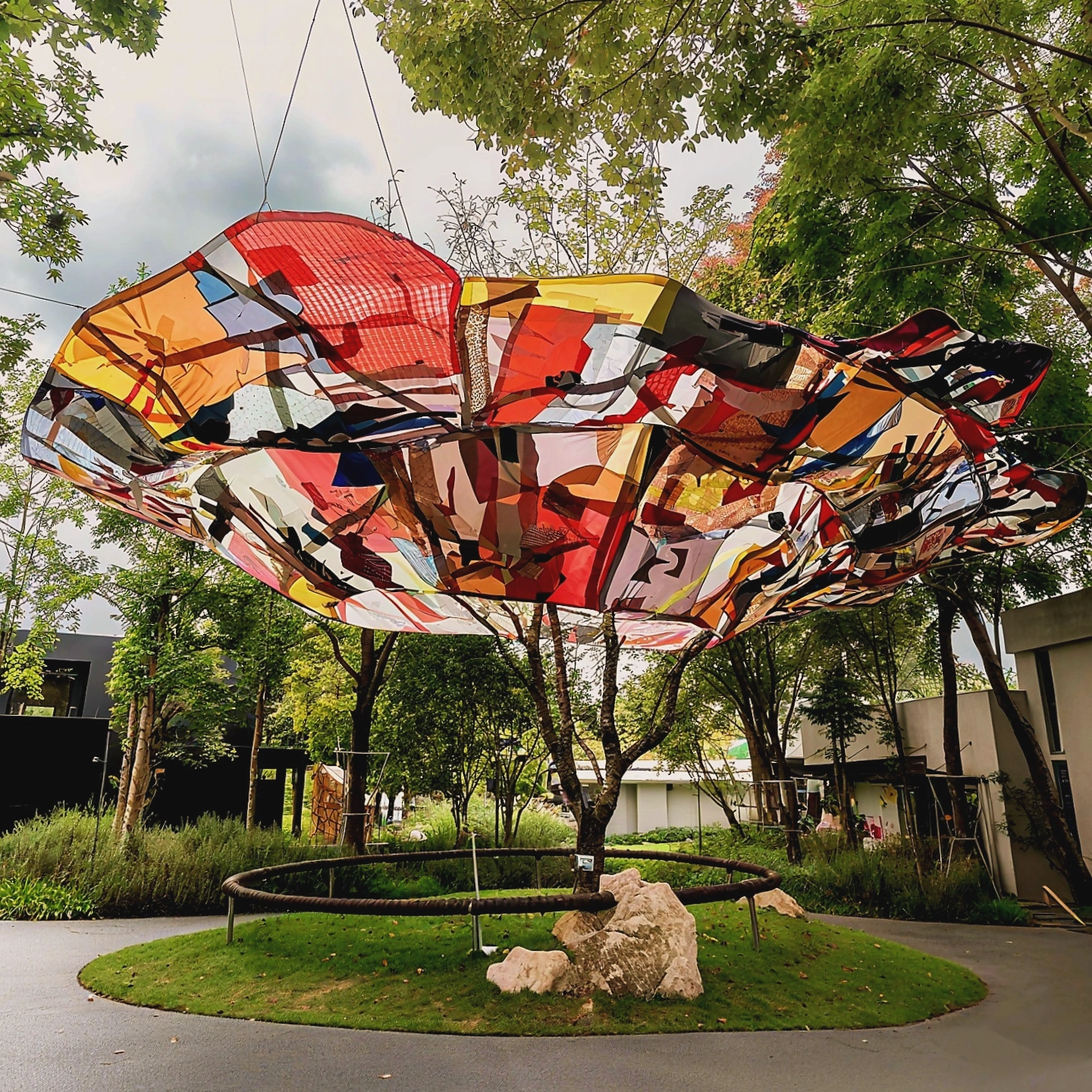 Colorful abstract sculpture suspended above a tree in a park, surrounded by greenery and pathways.