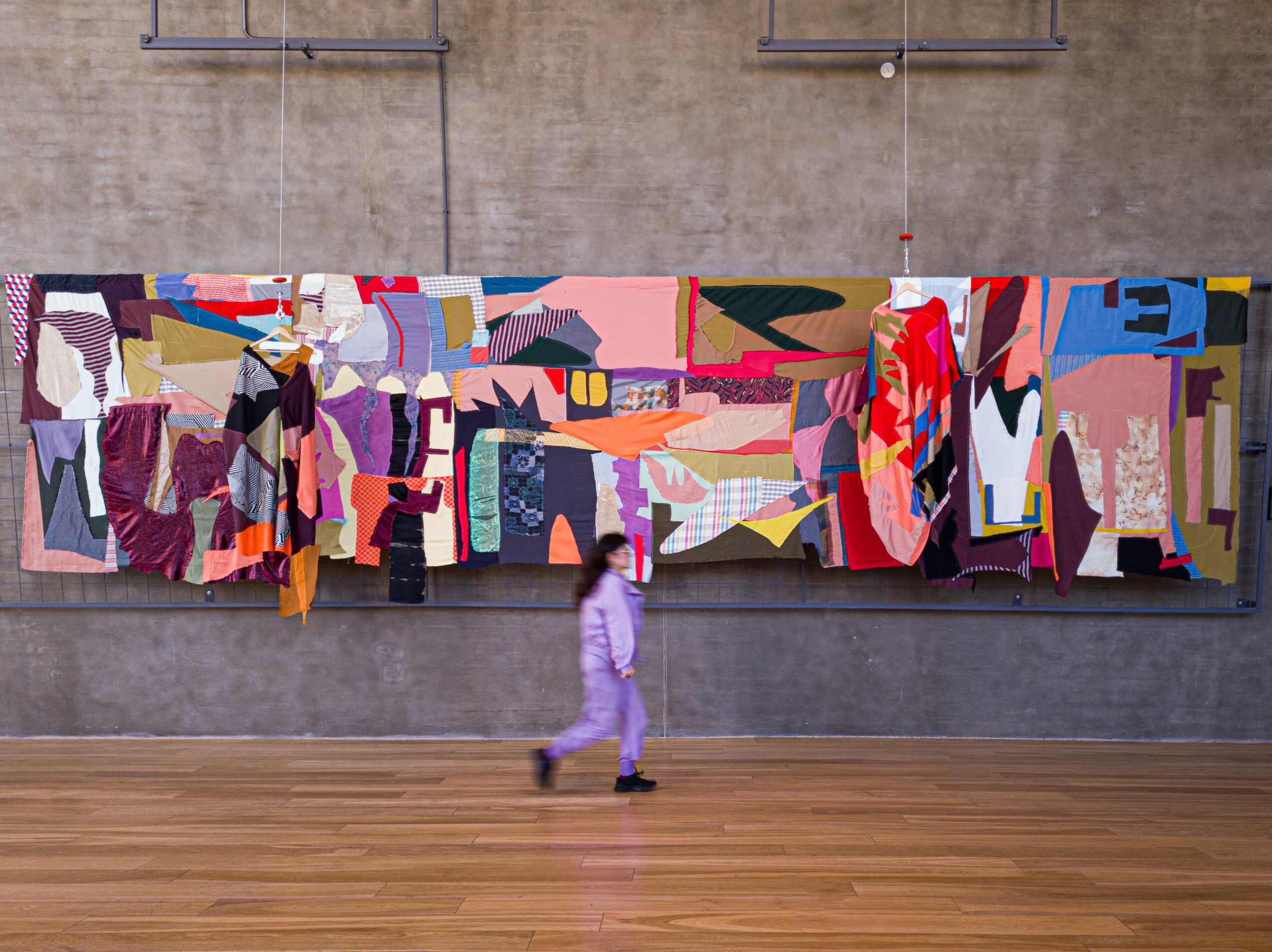 Person walking in front of a large colorful abstract artwork on a wall, with a wooden floor and concrete wall background.