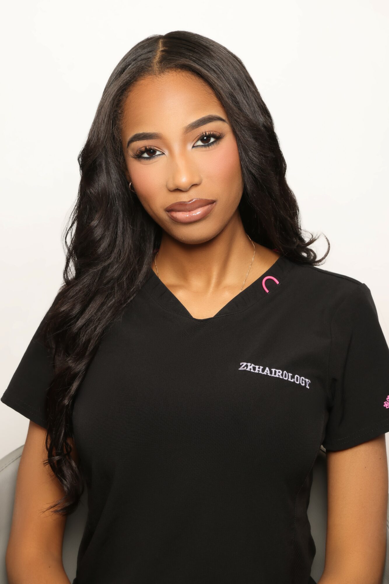 Young woman with long wavy hair wearing a black medical scrub top, looking at the camera with a neutral expression.