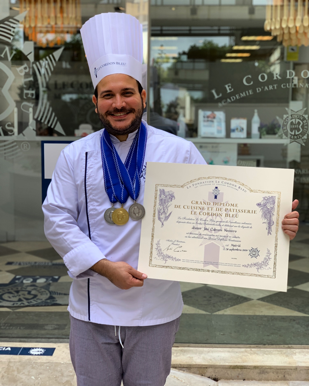 Chef wearing a tall white hat and medals, holding a certificate outside a building with glass windows.