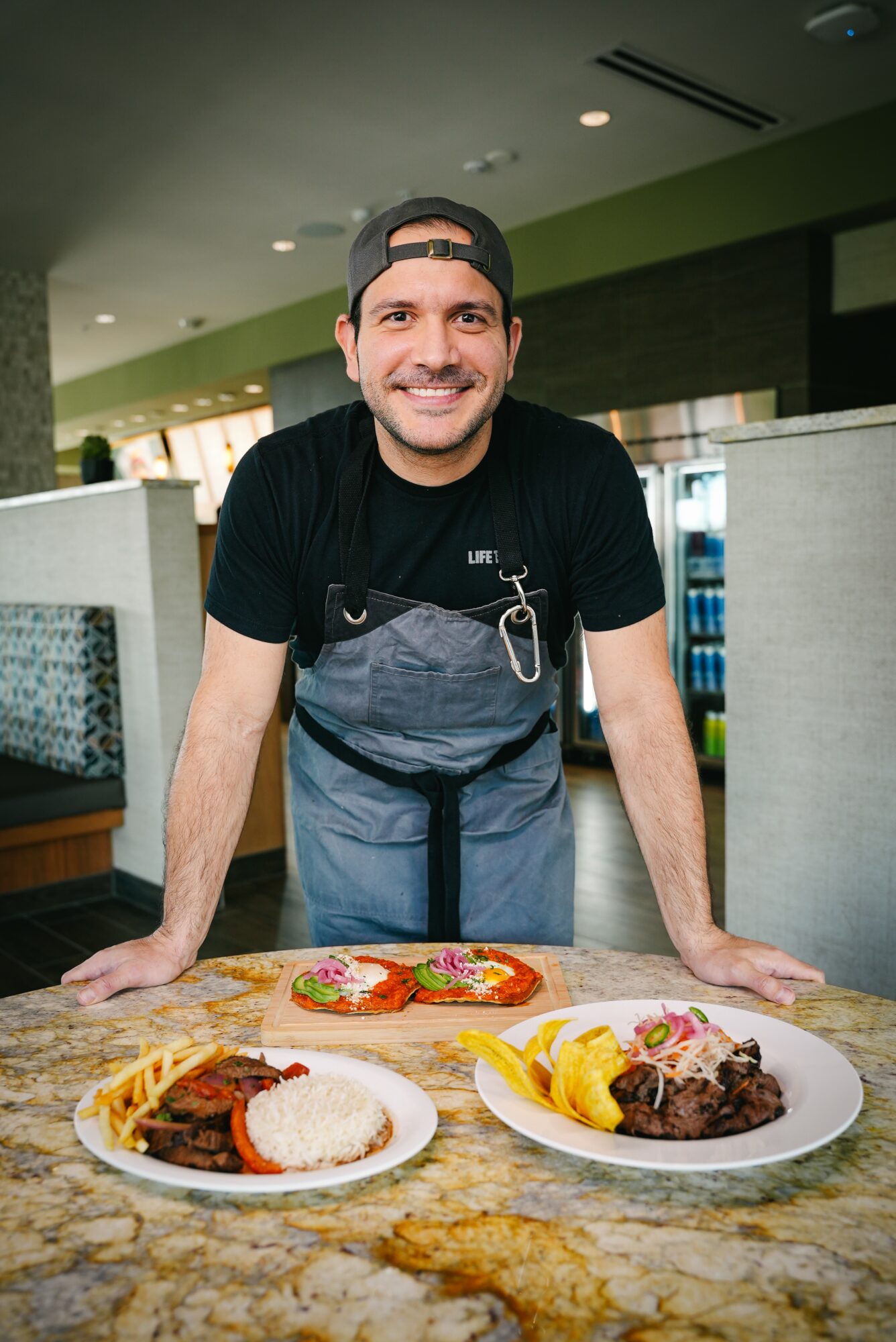Man smiling at camera, leaning on a counter with two plates of food, in a restaurant setting.