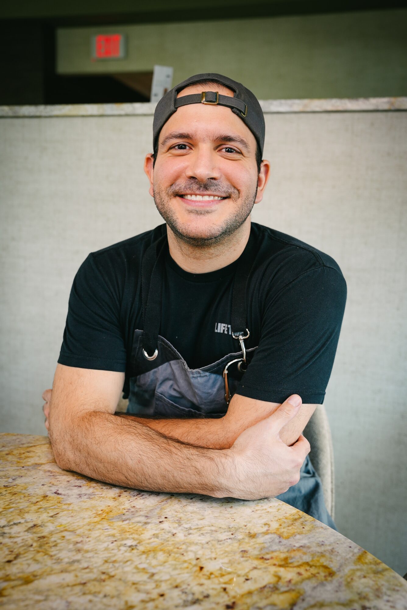 Smiling man with a beard, wearing a black cap backwards and black t-shirt, sitting at a table with arms crossed.