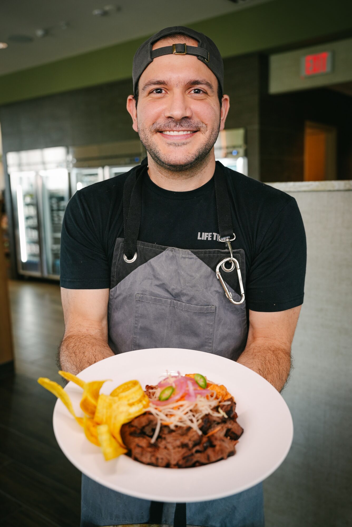 Man in black shirt and apron holding a plate of food, smiling, in a restaurant setting.