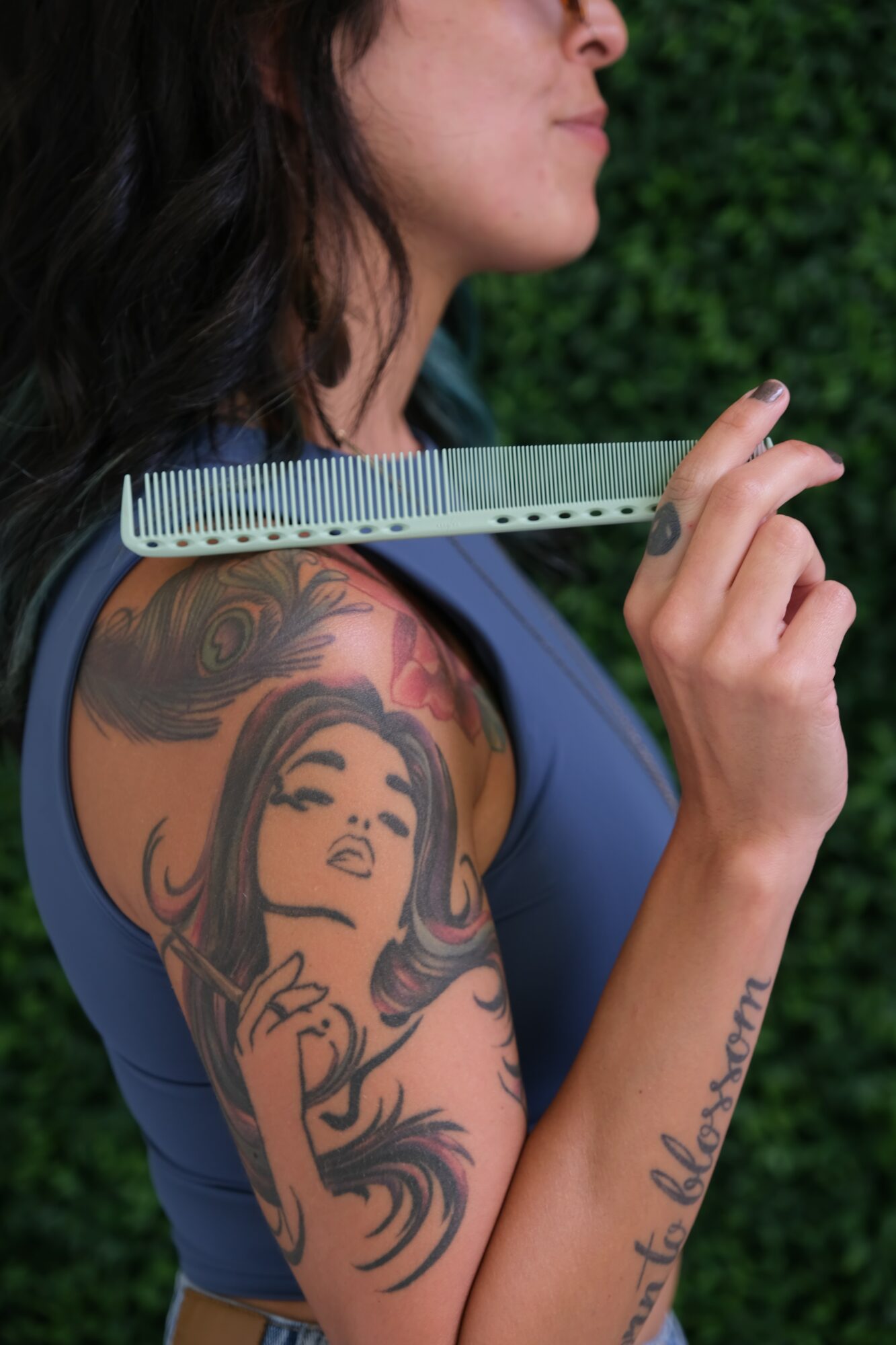 Close-up of a woman with dark hair and a tattooed arm holding a ruler near her face, outdoors with green foliage.
