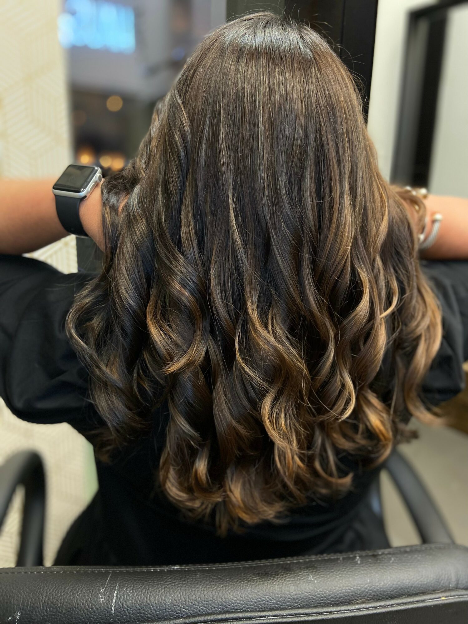 Back of a person with long, wavy brown hair, sitting in a chair indoors, with a blurred background.
