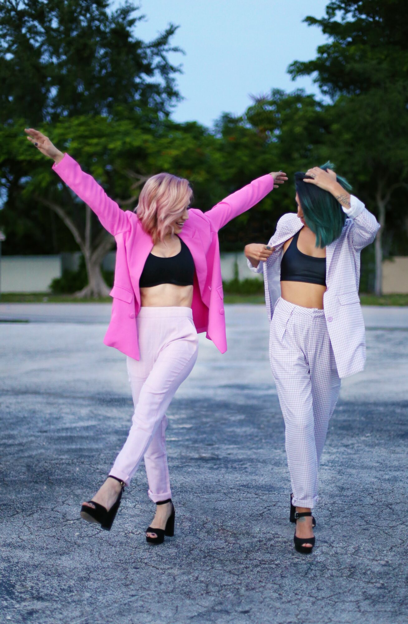 Two women in casual outfits pose outdoors with arms raised, standing on a paved surface with trees in the background.