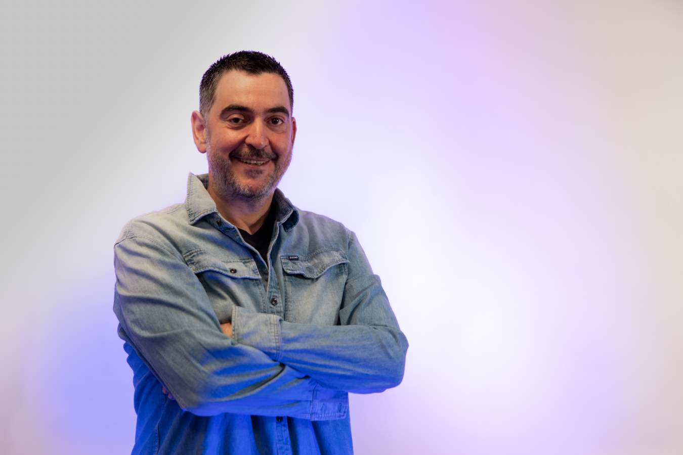 Smiling man with crossed arms, wearing a denim jacket, standing against a plain light background.