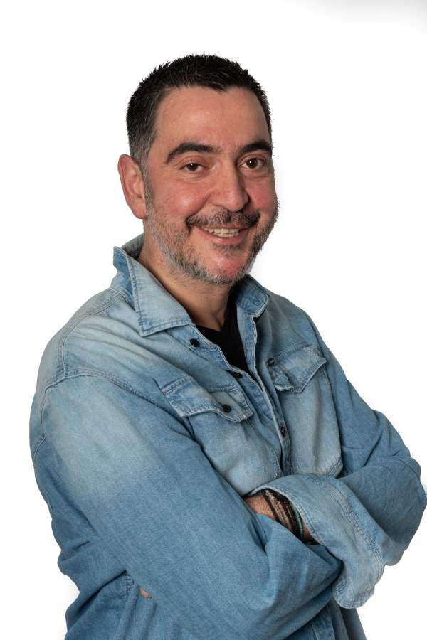 Smiling man with short dark hair and beard, wearing a denim jacket, arms crossed, against a white background.