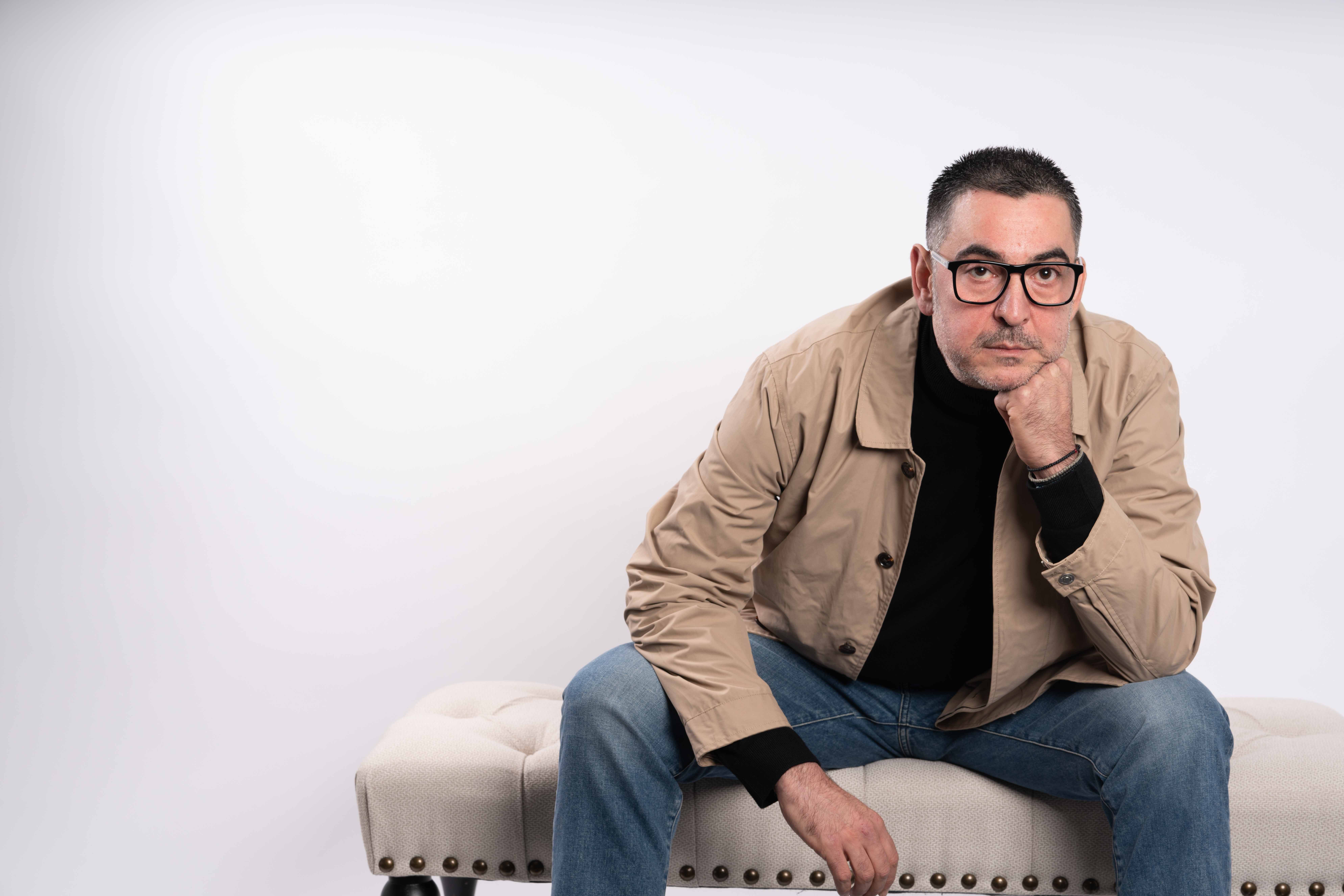 Man with glasses sitting on a beige bench, resting his chin on his hand, against a plain white background.