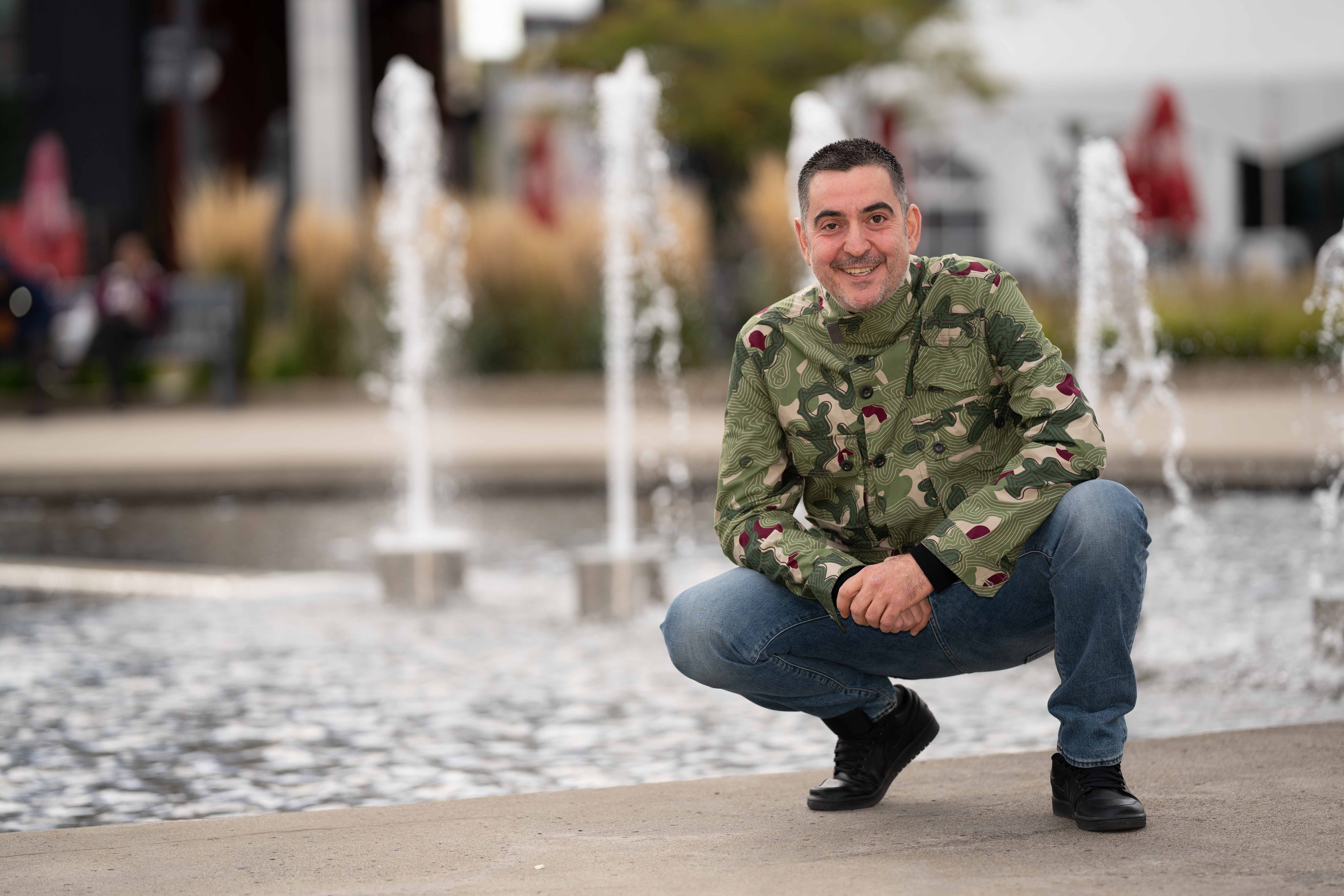 Man crouching near a fountain, smiling, wearing a camouflage jacket and jeans, in an outdoor setting with water jets in the background.