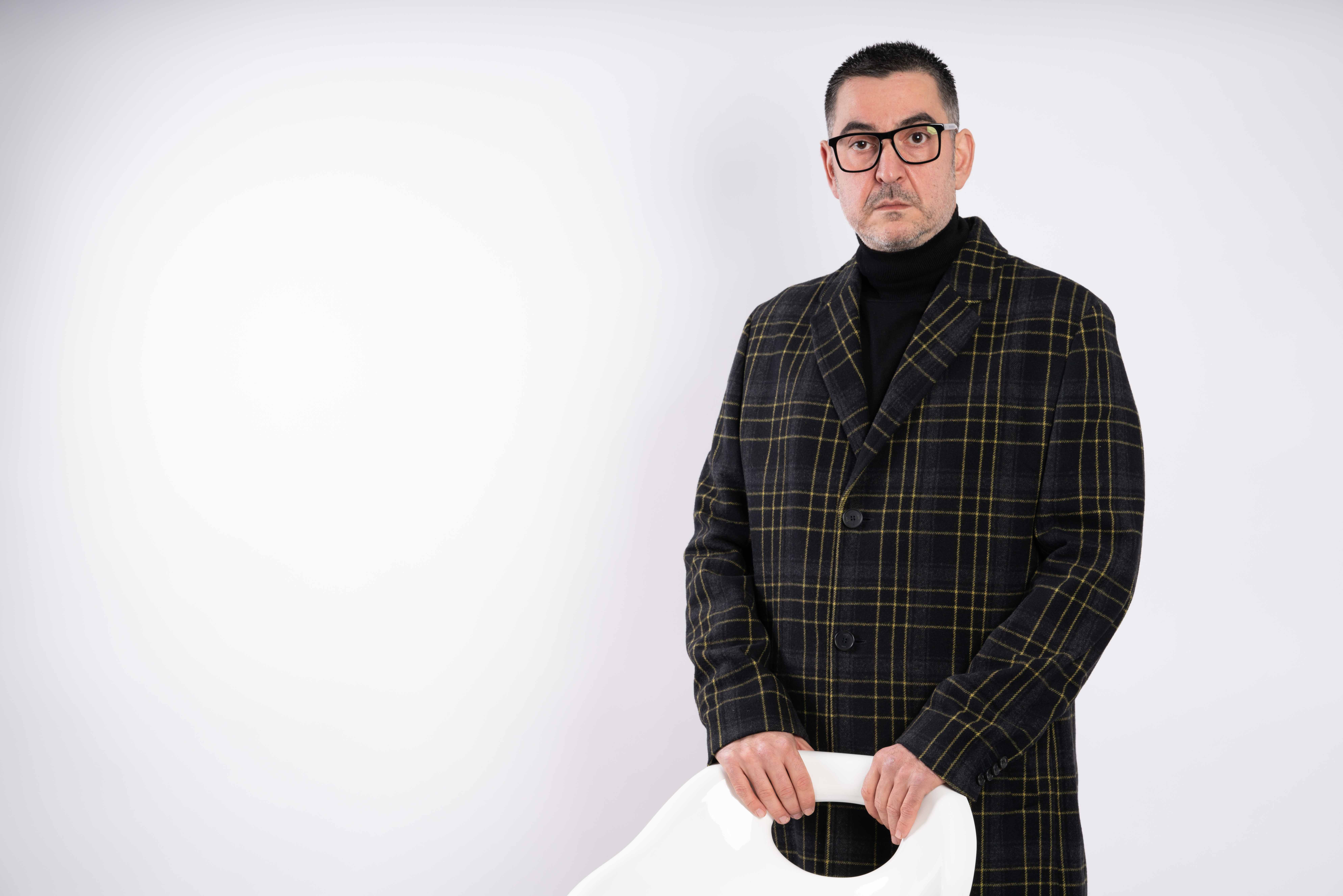Man with glasses wearing a dark plaid jacket and black turtleneck, standing against a plain white background.