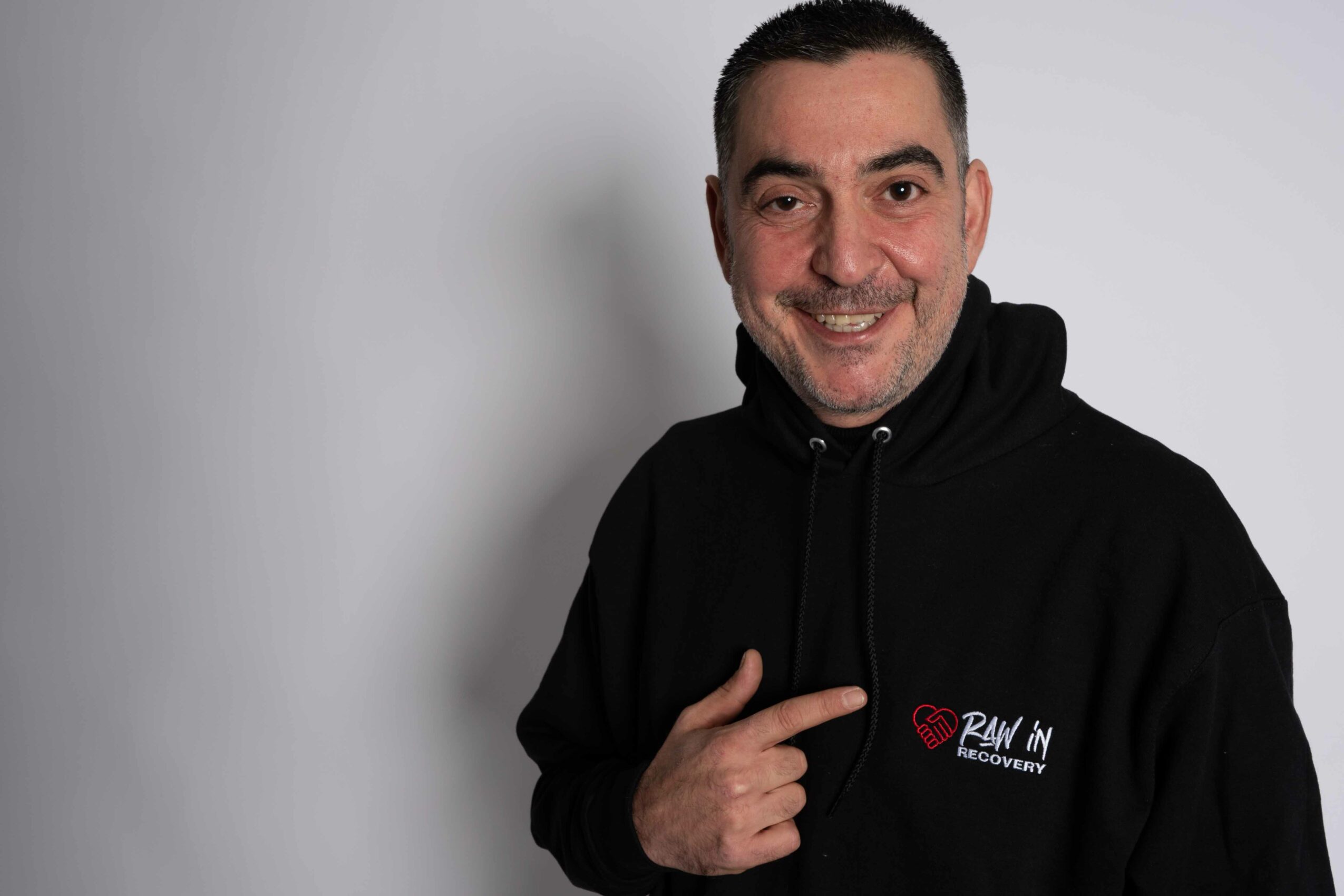 Man with short dark hair and a beard, smiling, wearing a black hoodie, pointing to the hoodie logo, plain background.