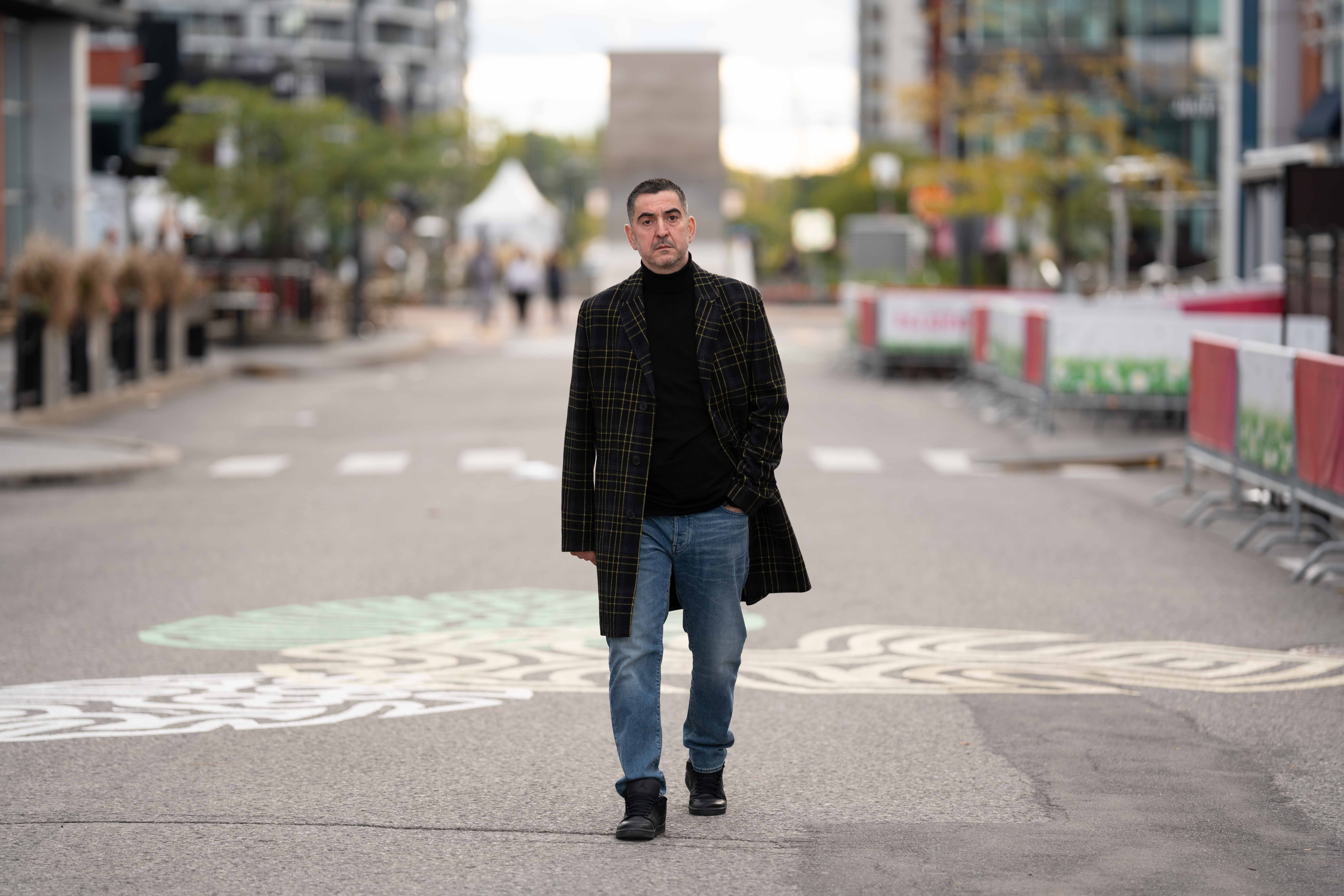 Man walking on city street with buildings and trees in background, wearing dark coat and jeans.