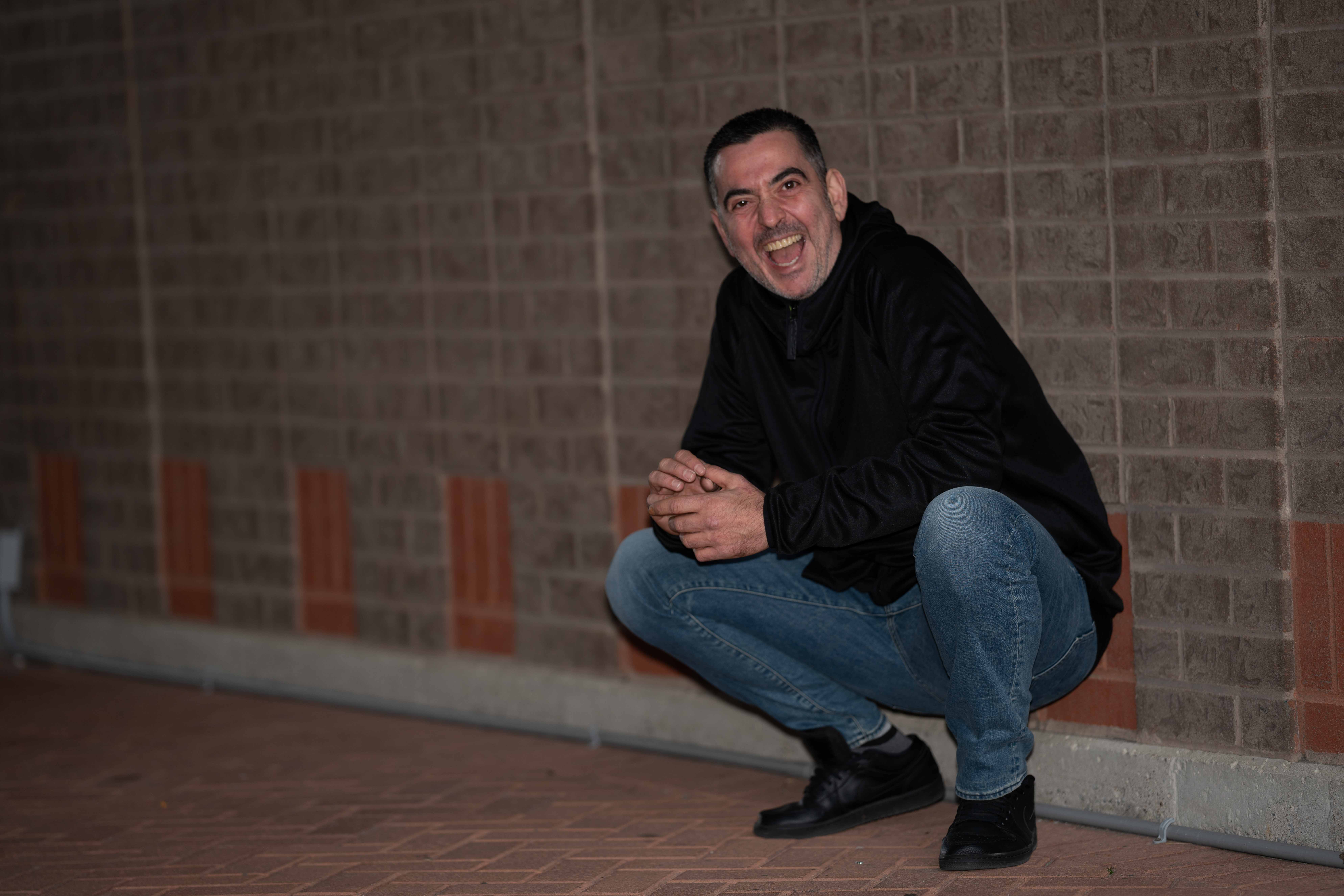 Man crouching and smiling against a brick wall, wearing a black jacket and blue jeans.