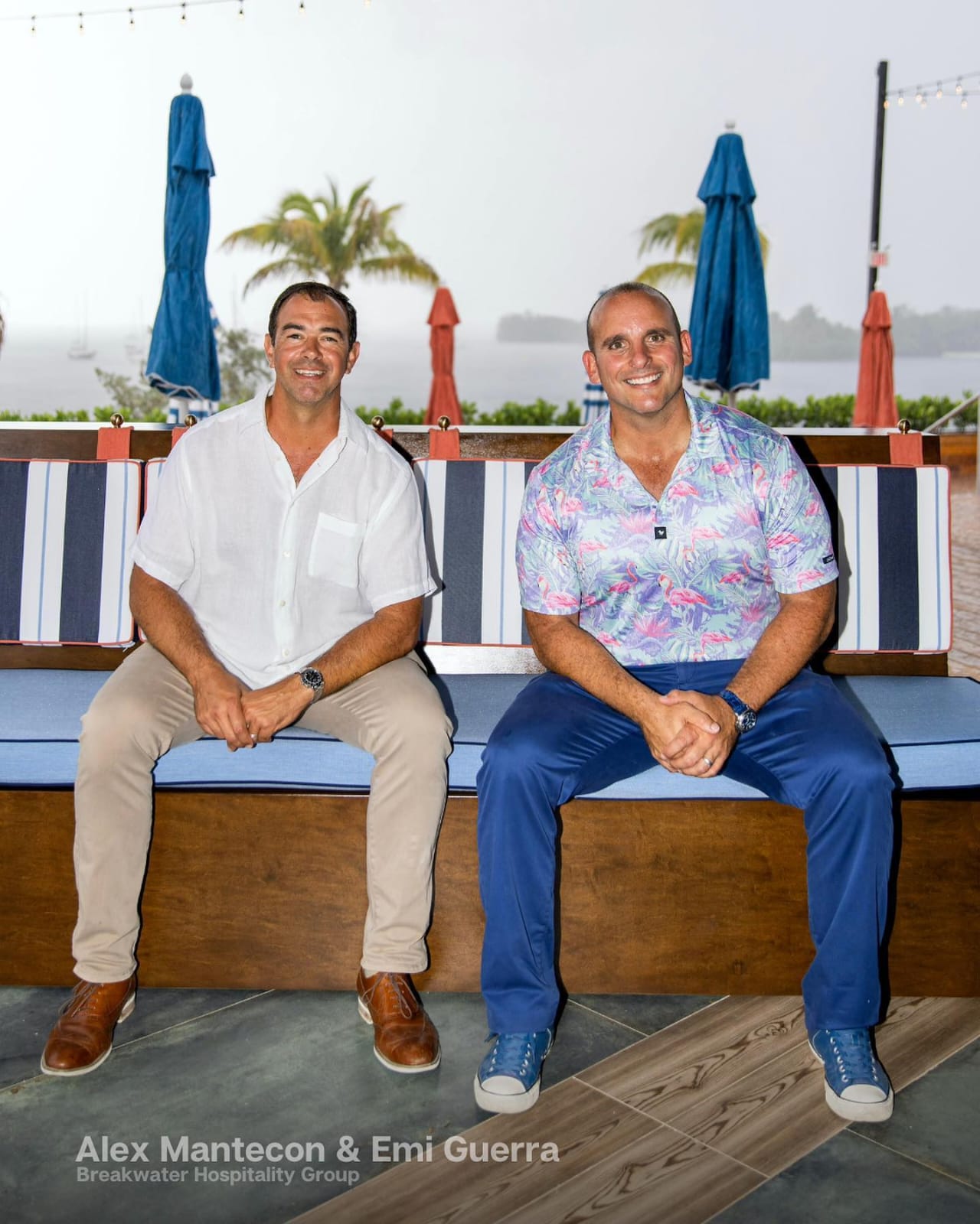 Two men sit on a bench outdoors with umbrellas and palm trees in the background, smiling at the camera.