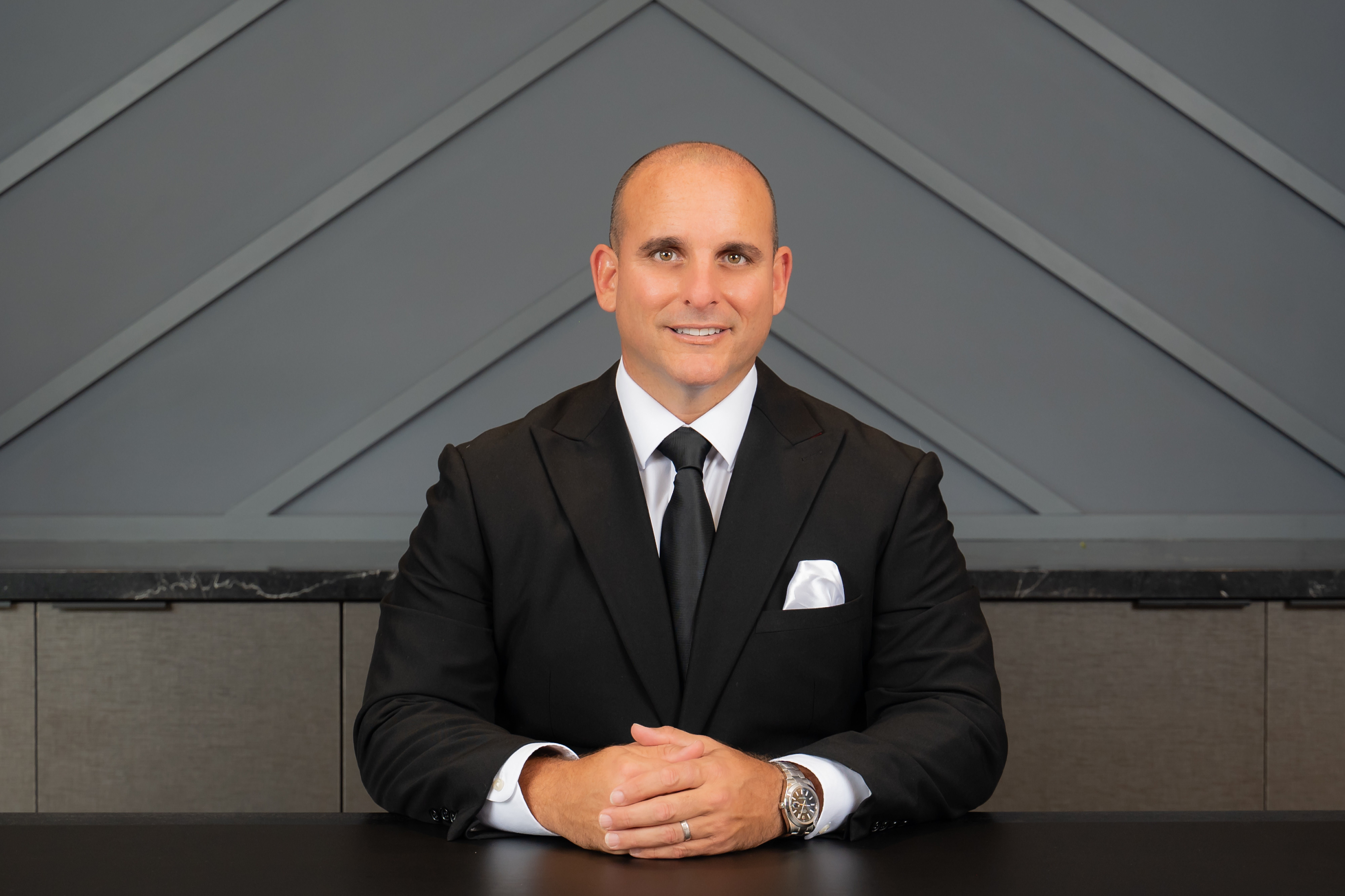 Man in a black suit and tie sitting at a table with hands clasped, smiling, in front of a geometric wall background.