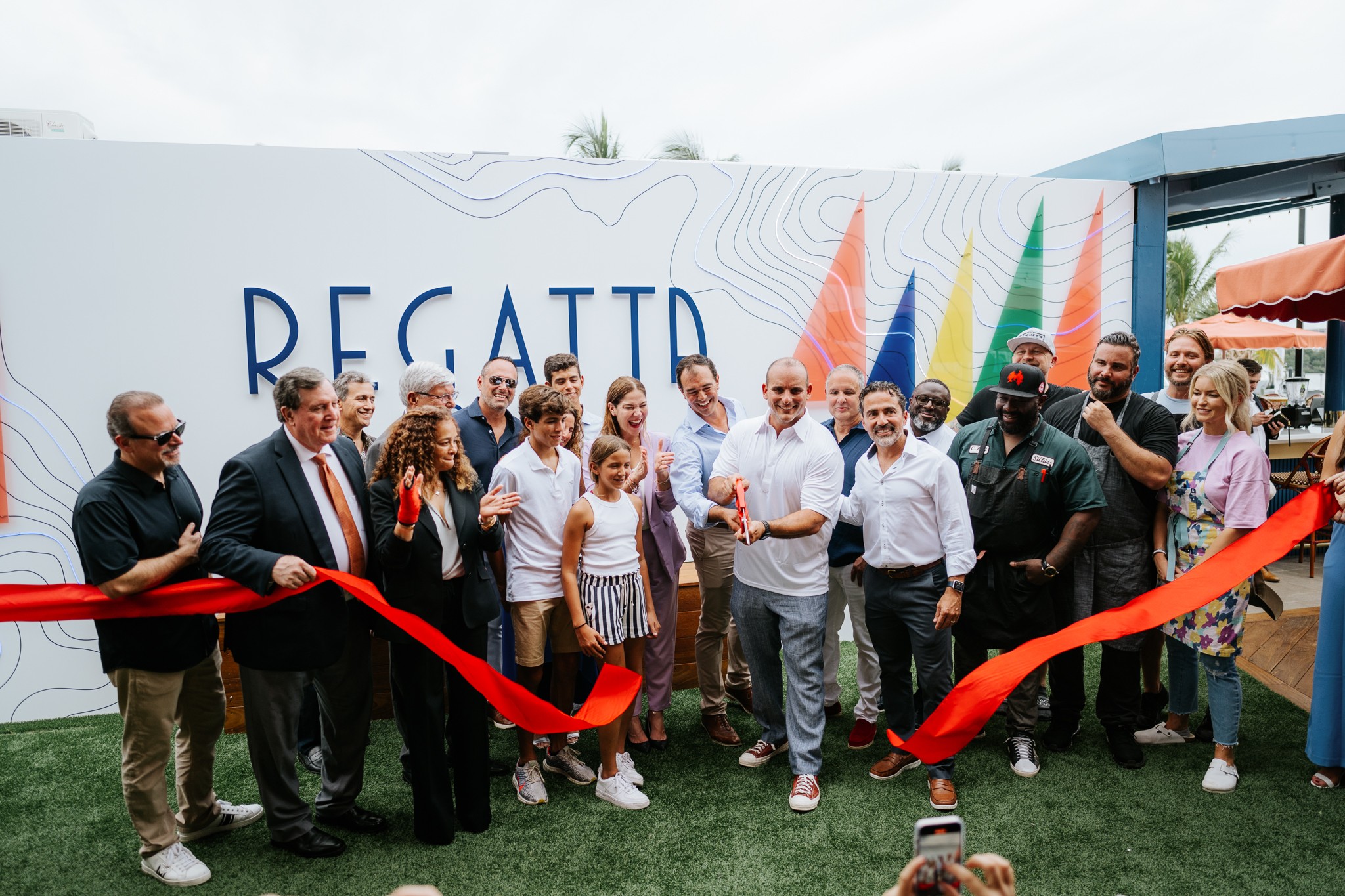 Group of people at a ribbon-cutting event in front of a sign that reads 'REGAITTO,' with some holding a red ribbon and others taking photos.