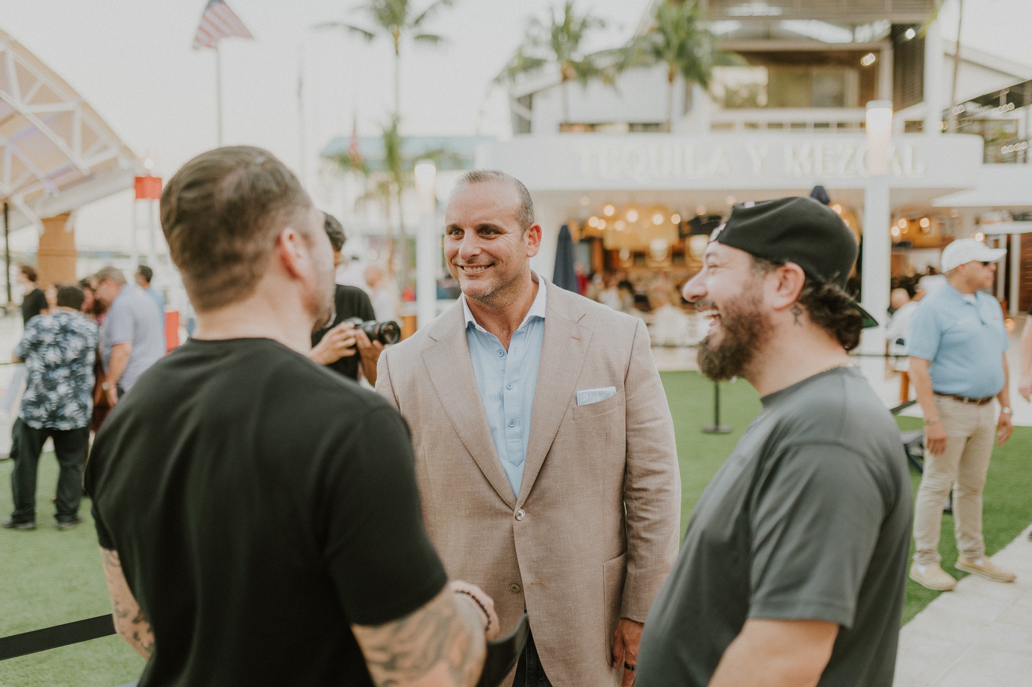 Three men engaged in conversation outdoors at an event, with a background of people, tents, and palm trees.