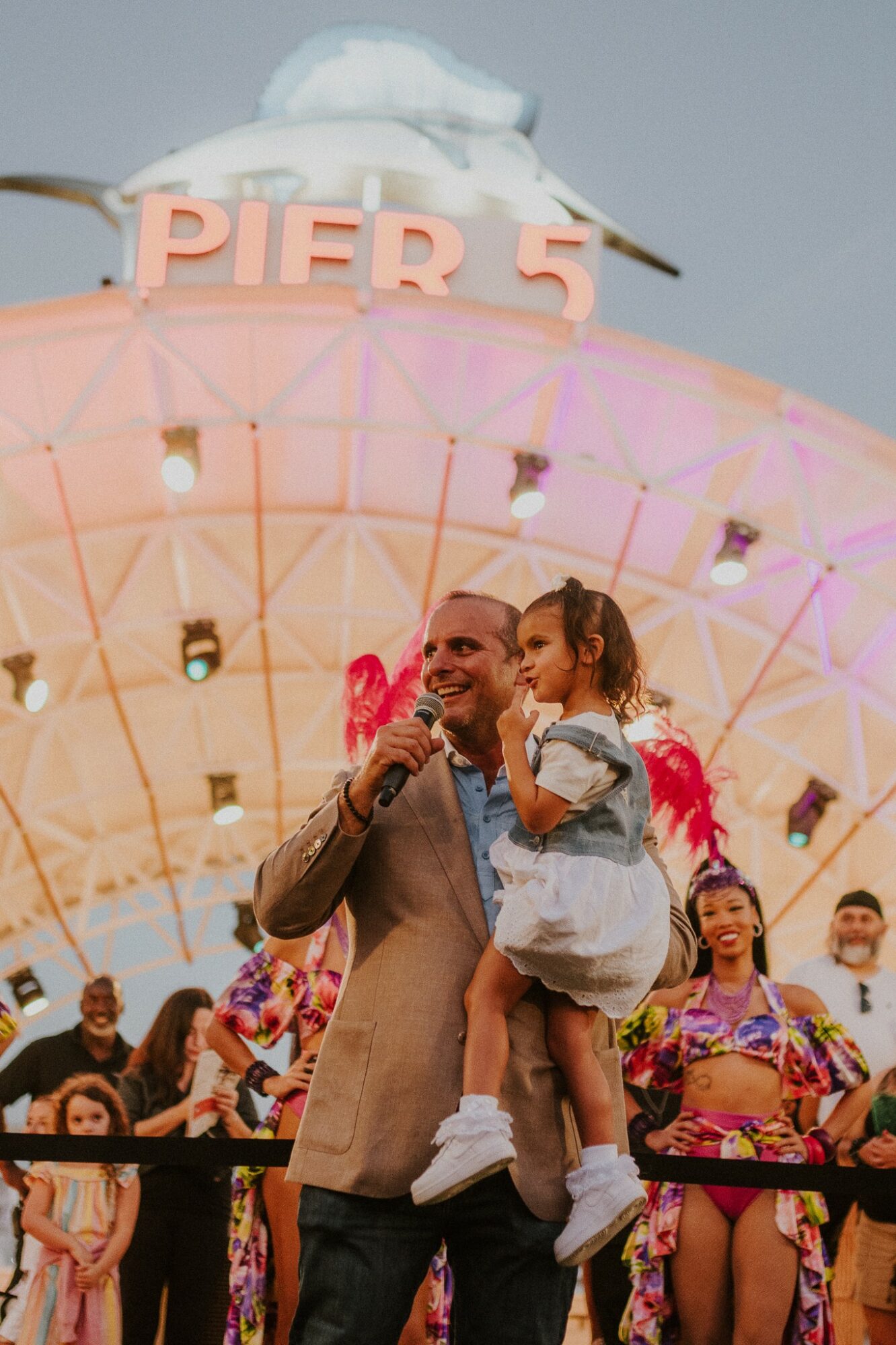 A man holding a young girl at Pier 5 amusement park with a large Ferris wheel in the background, colorful lights, and other people around.