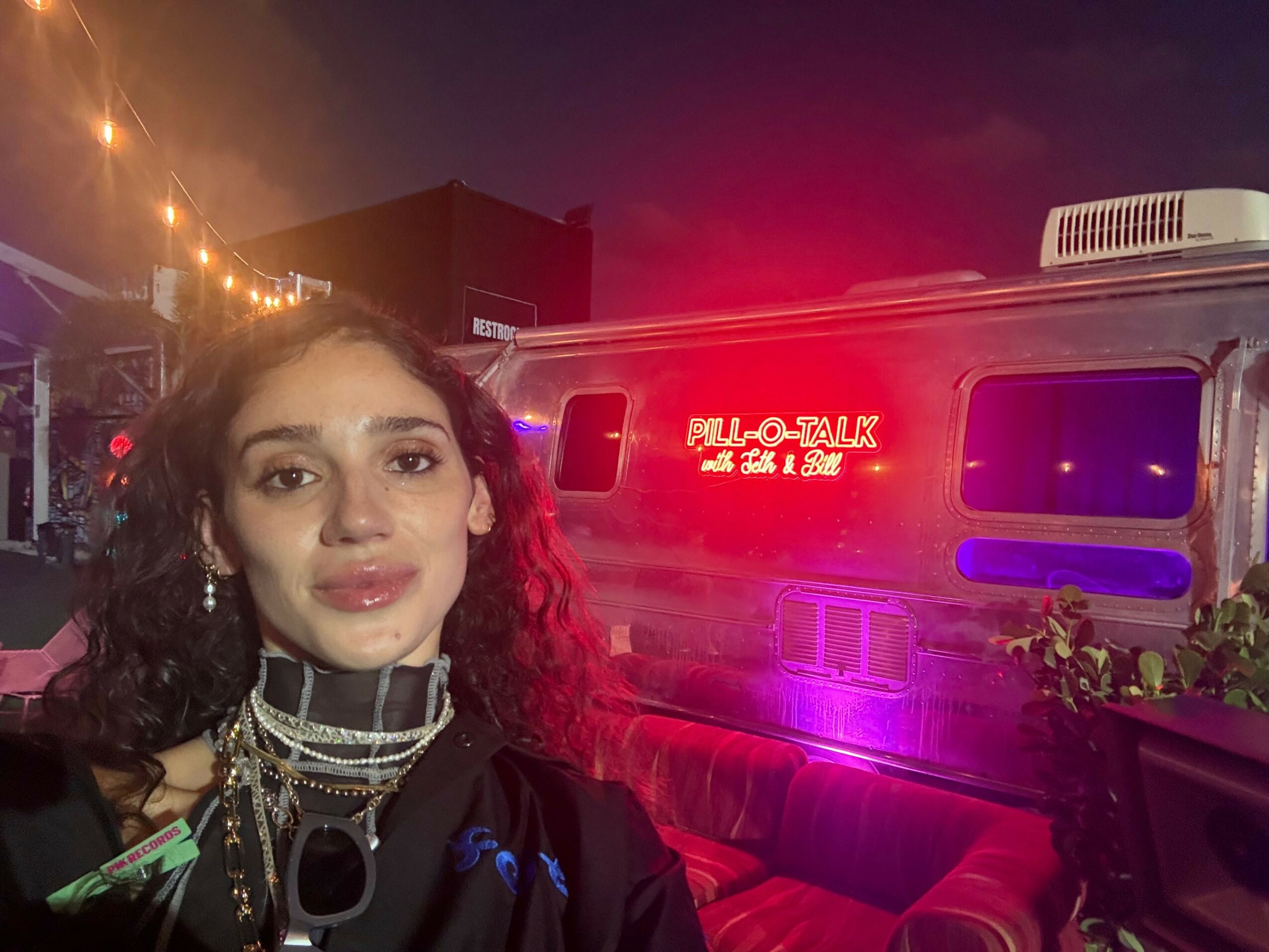 Upside-down woman with dark hair and makeup, wearing a black and silver outfit, standing outdoors at night with lights and a building in the background.