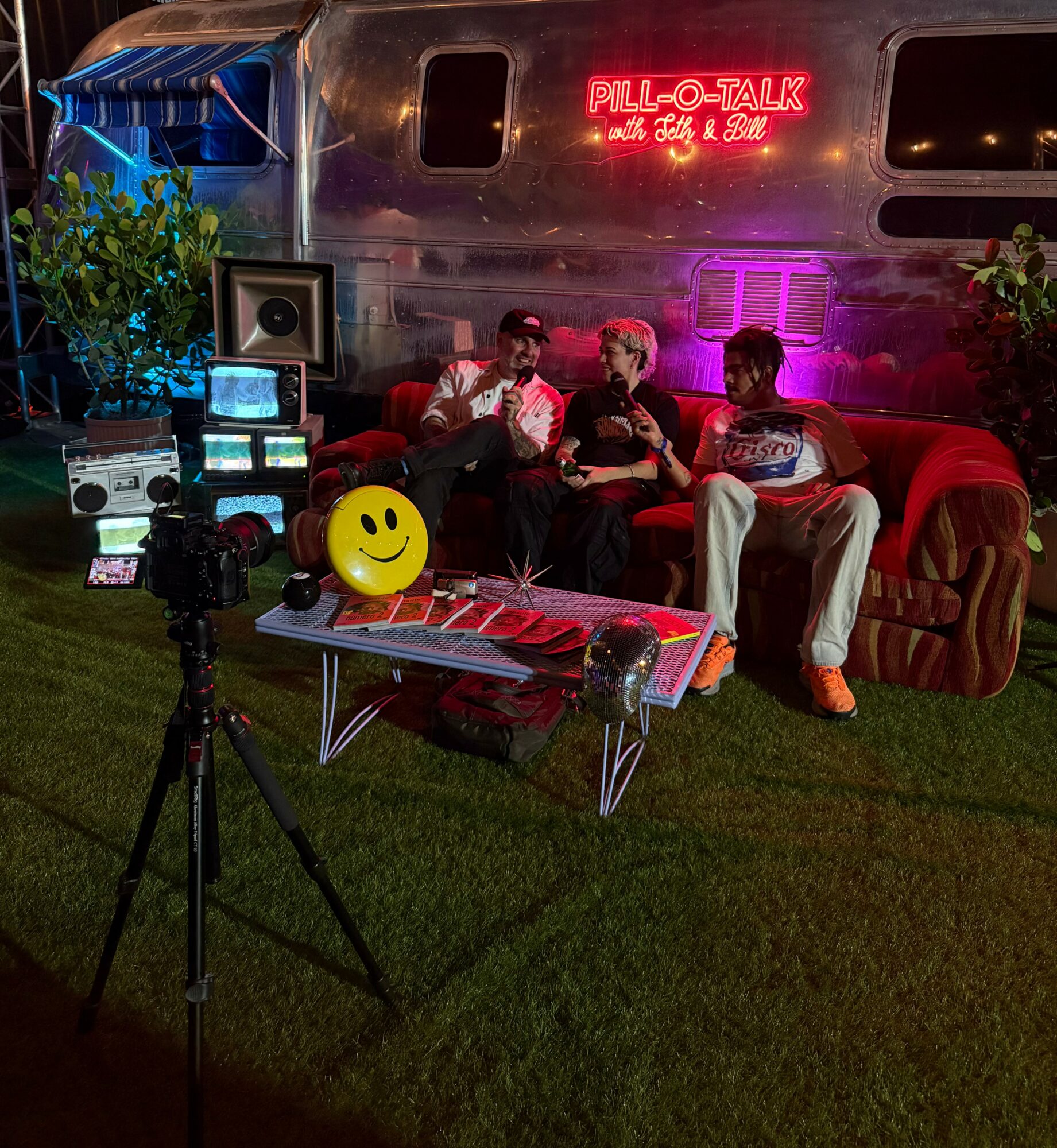 Three people sit on a red sofa outdoors at night, with a neon sign, plants, and equipment nearby.