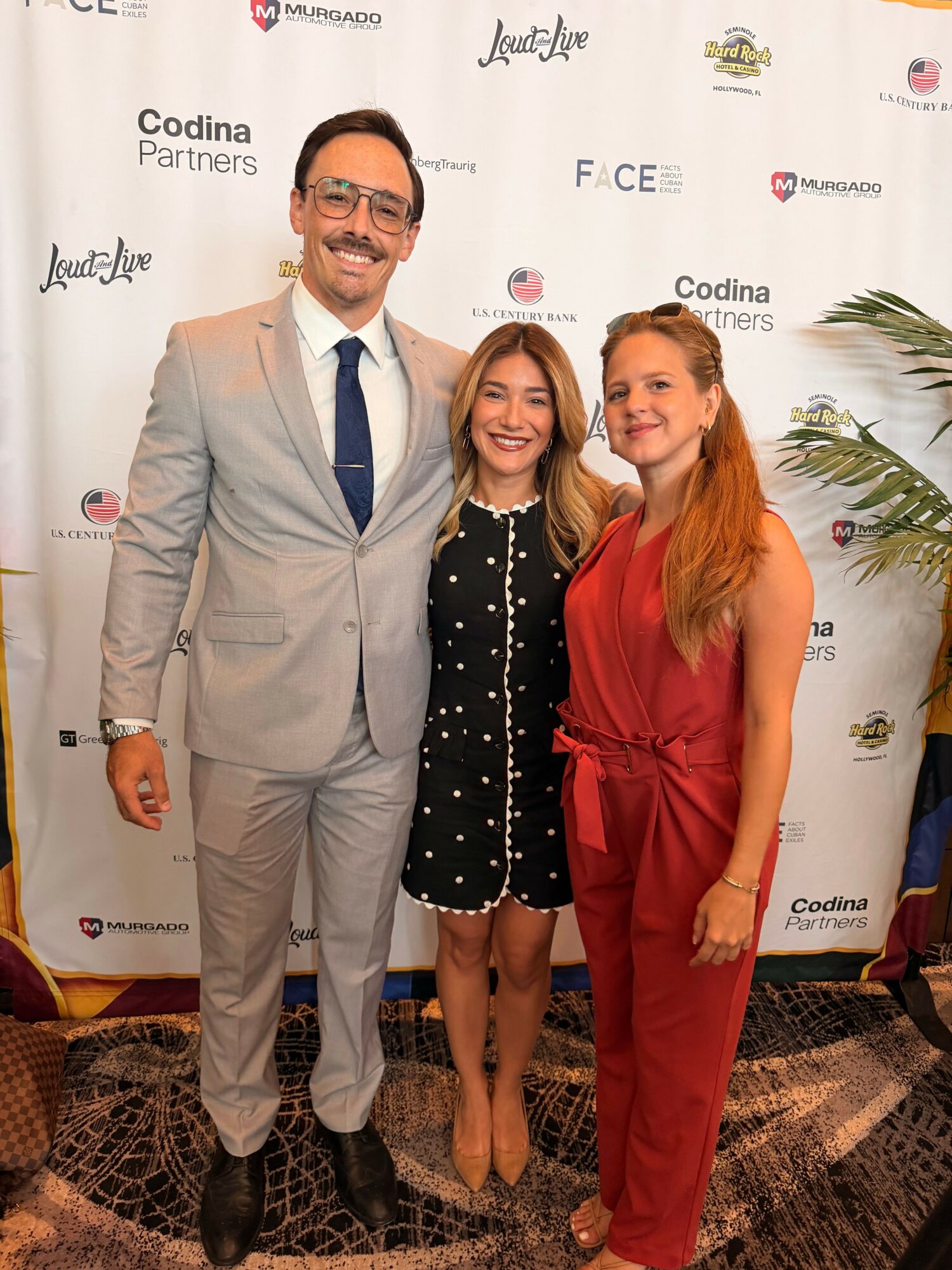 Three people standing together at an event, with a backdrop displaying logos and text, smiling for the photo.