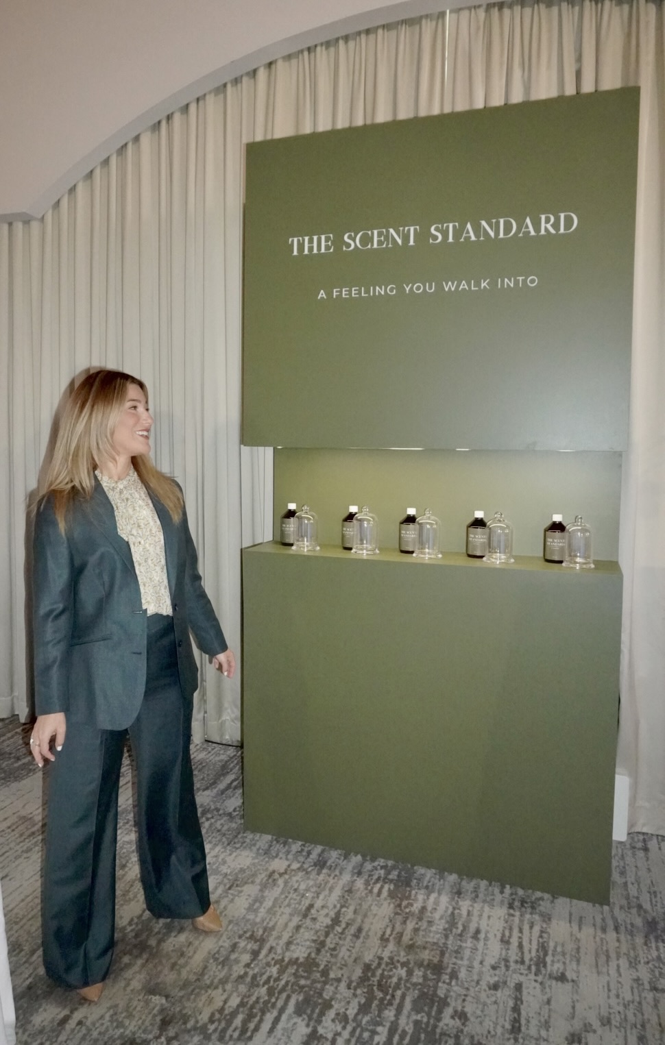 Woman in a suit looking at a display with bottles and a sign reading 'The Scent Standard' and 'A Feeling You Walk Into'.