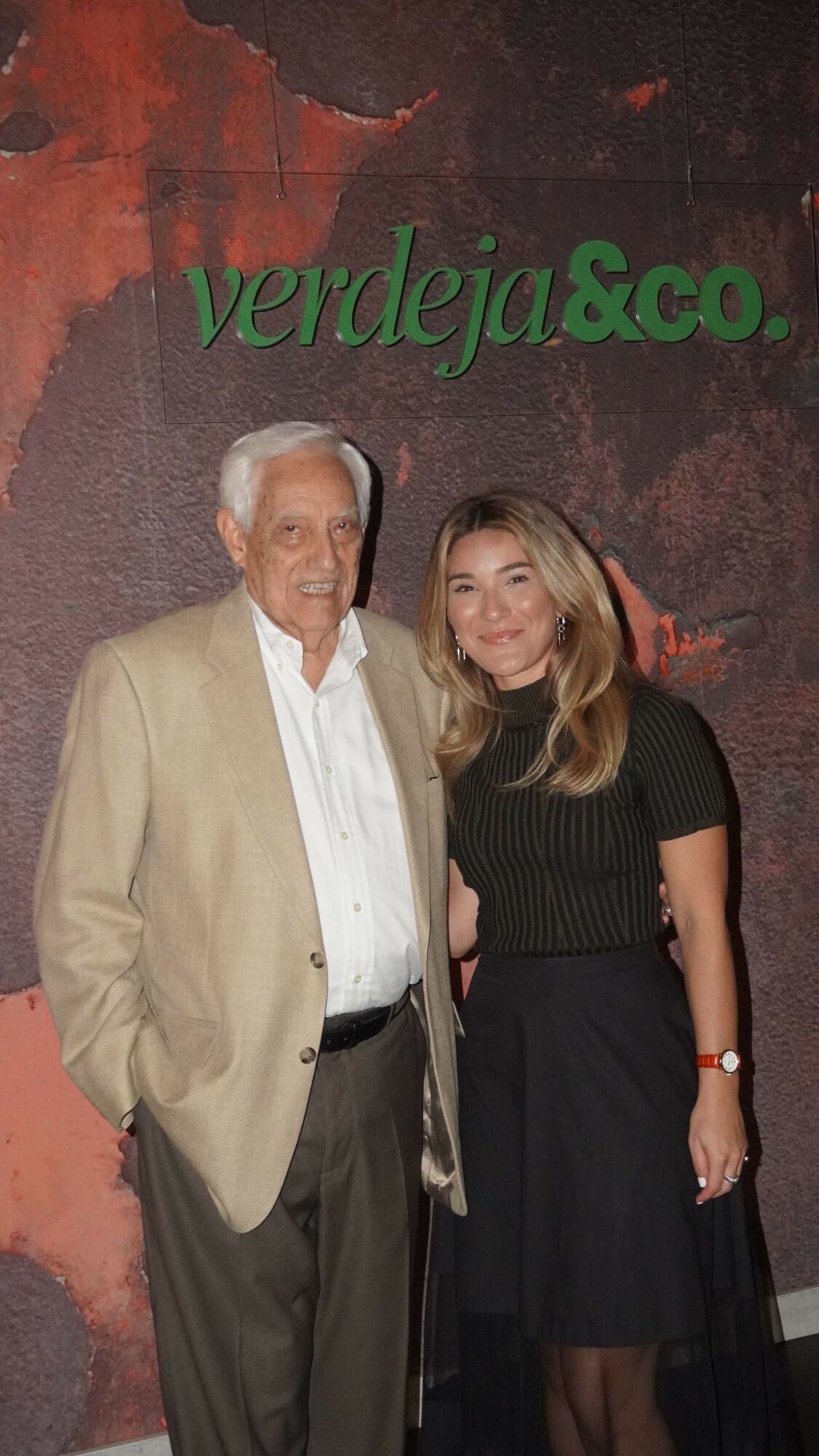 Two people standing side by side in front of a textured background with green text, one man and one woman, both smiling.