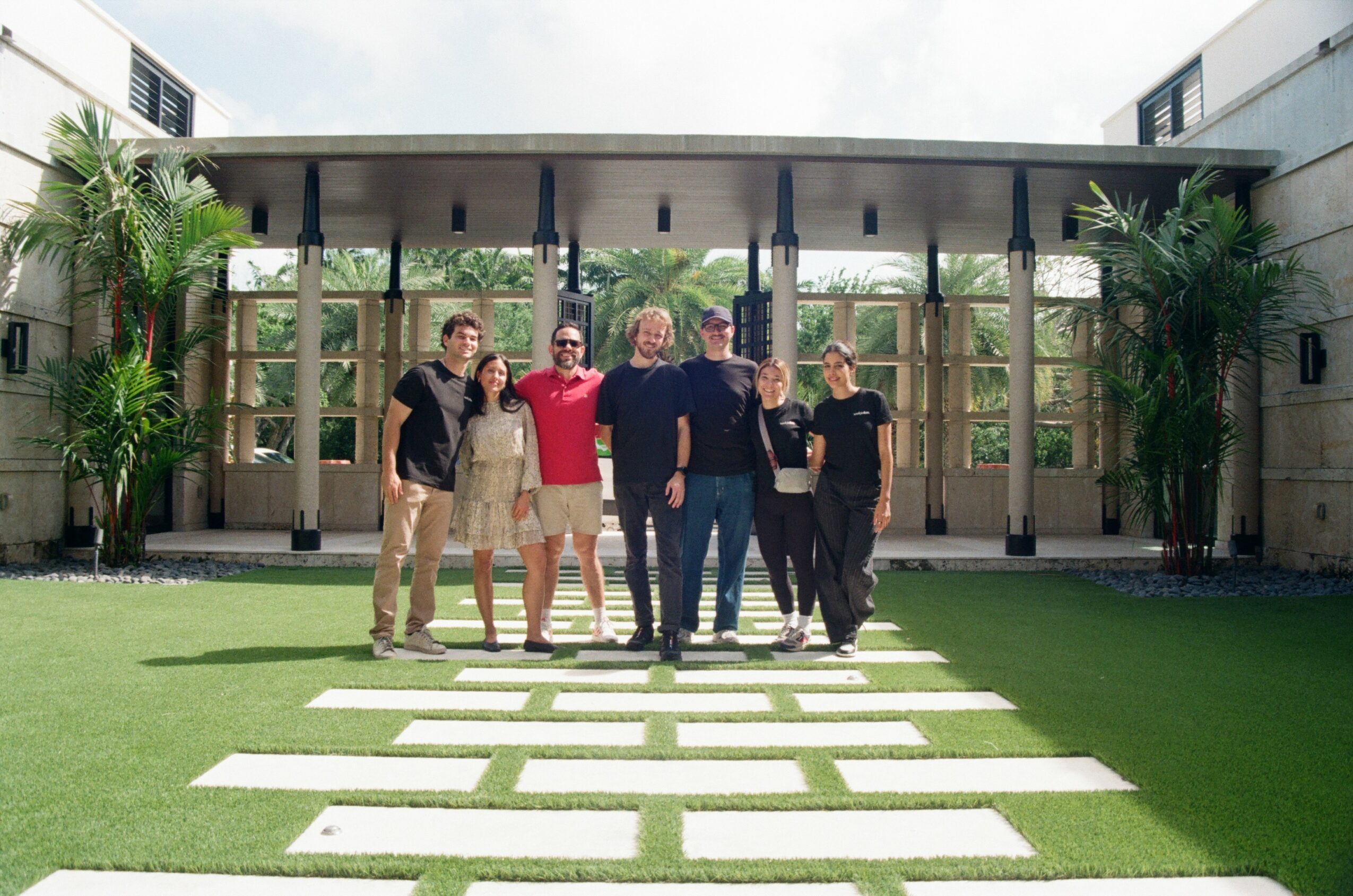 Group of six people standing on a pathway in front of a modern building with greenery, smiling outdoors.