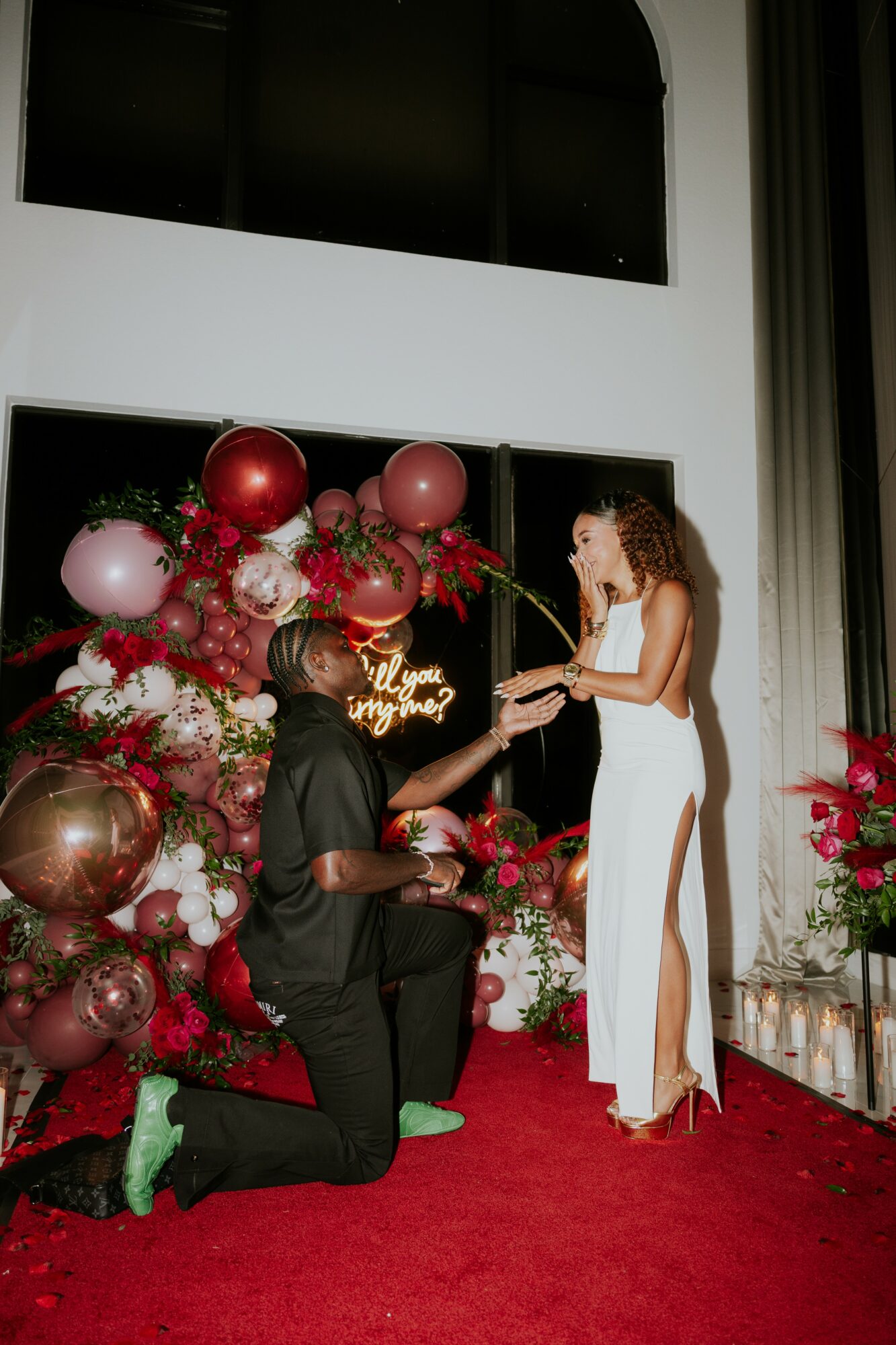 A woman in a white dress receives a ring from a man kneeling on a red carpet, surrounded by pink and red balloons and flowers.