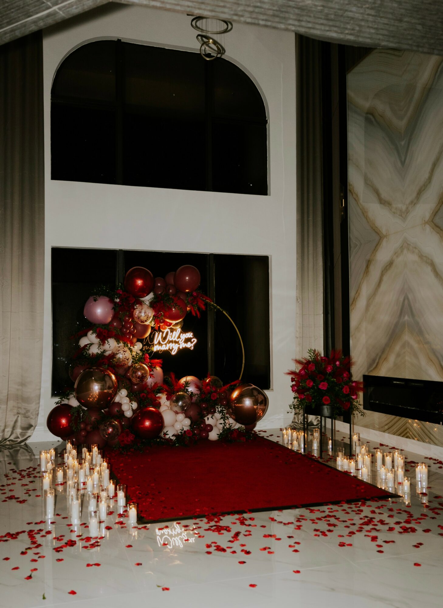 Decorated entrance with red and pink balloons, flowers, candles, and rose petals on a red carpet, in a modern interior.