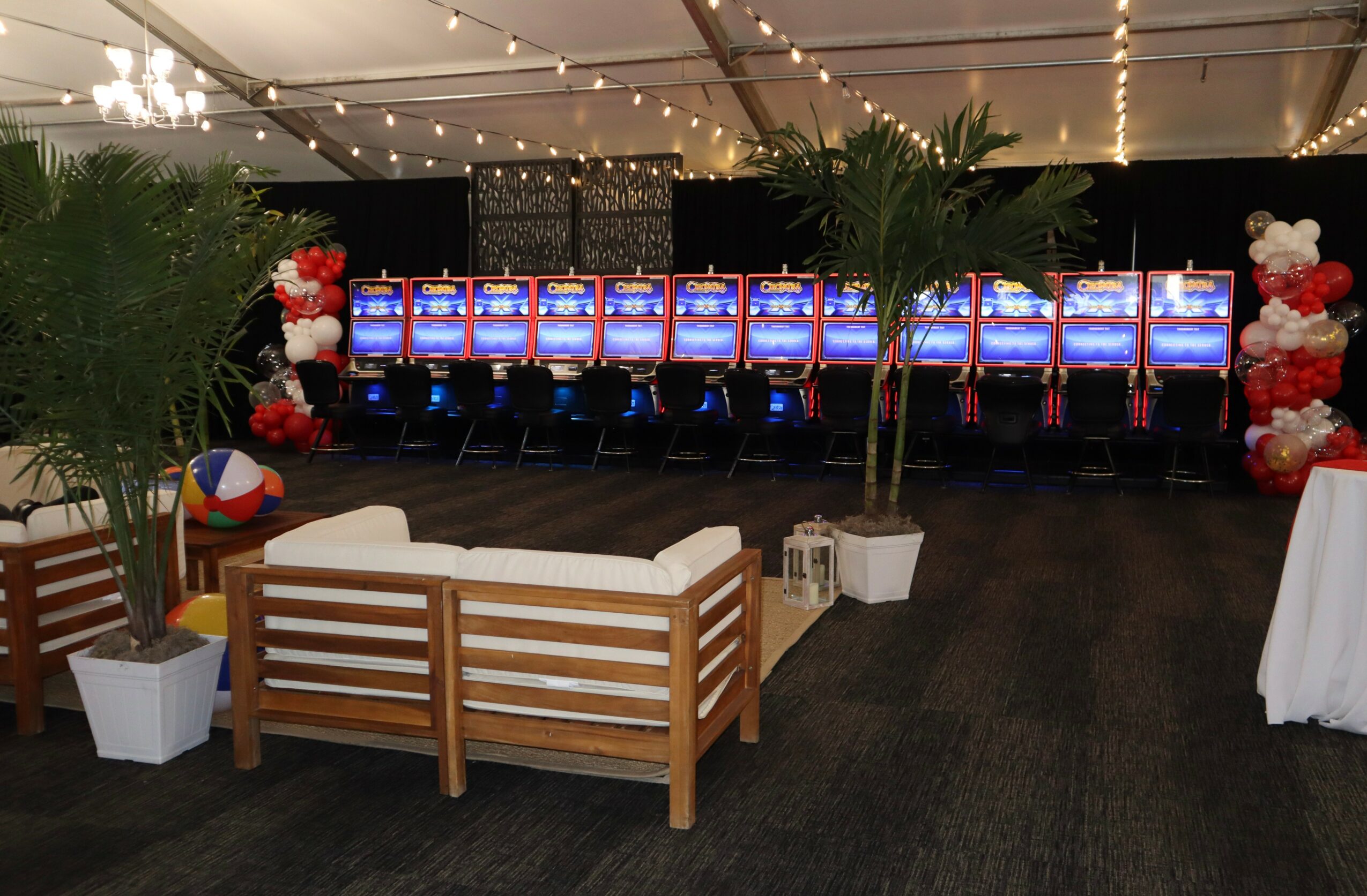 Casino gaming area with slot machines, a wooden bench, potted plants, and balloon decorations, under a tent ceiling.
