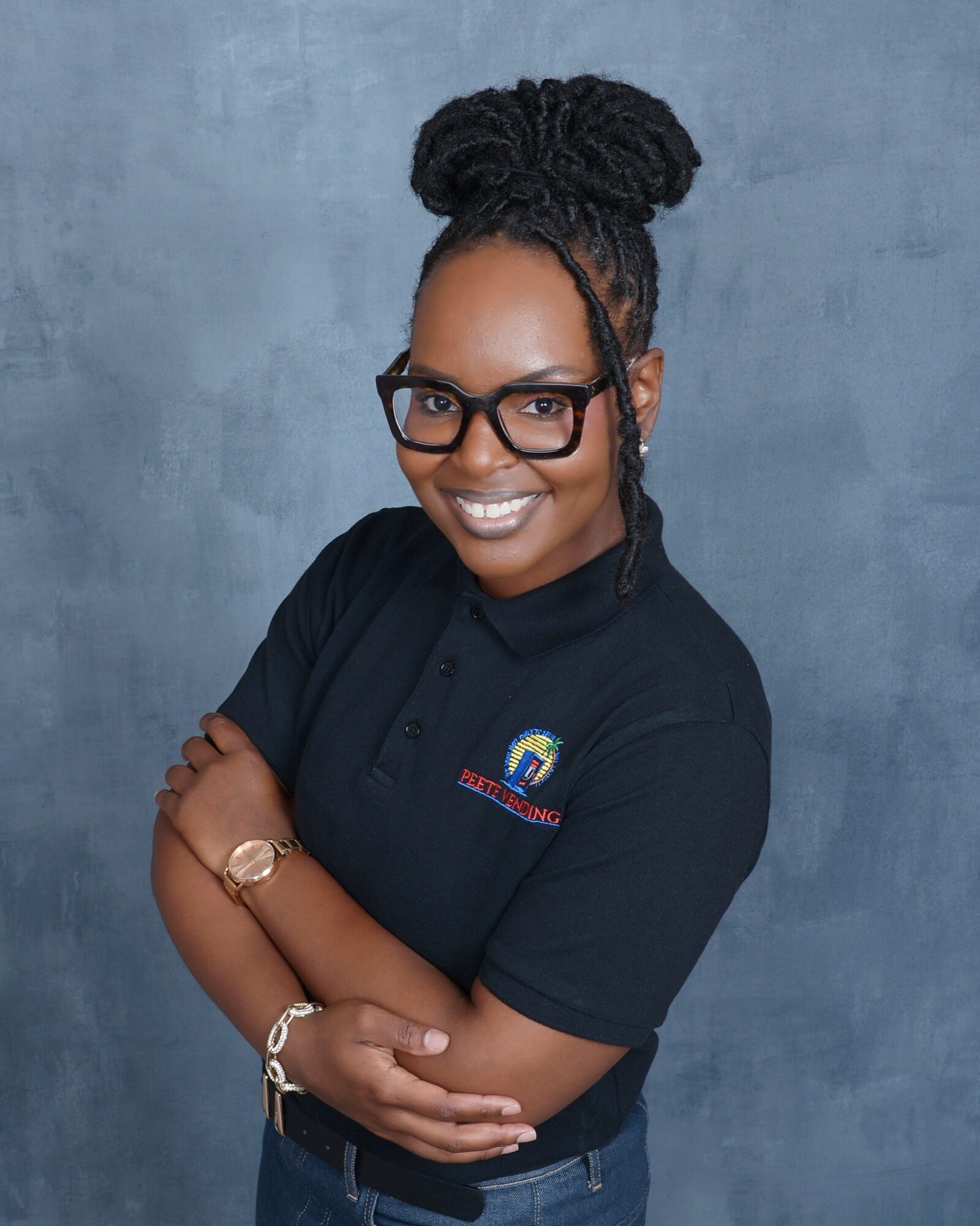Woman with glasses and braided hair styled in an updo, smiling, wearing a black polo shirt, standing against a gray background.