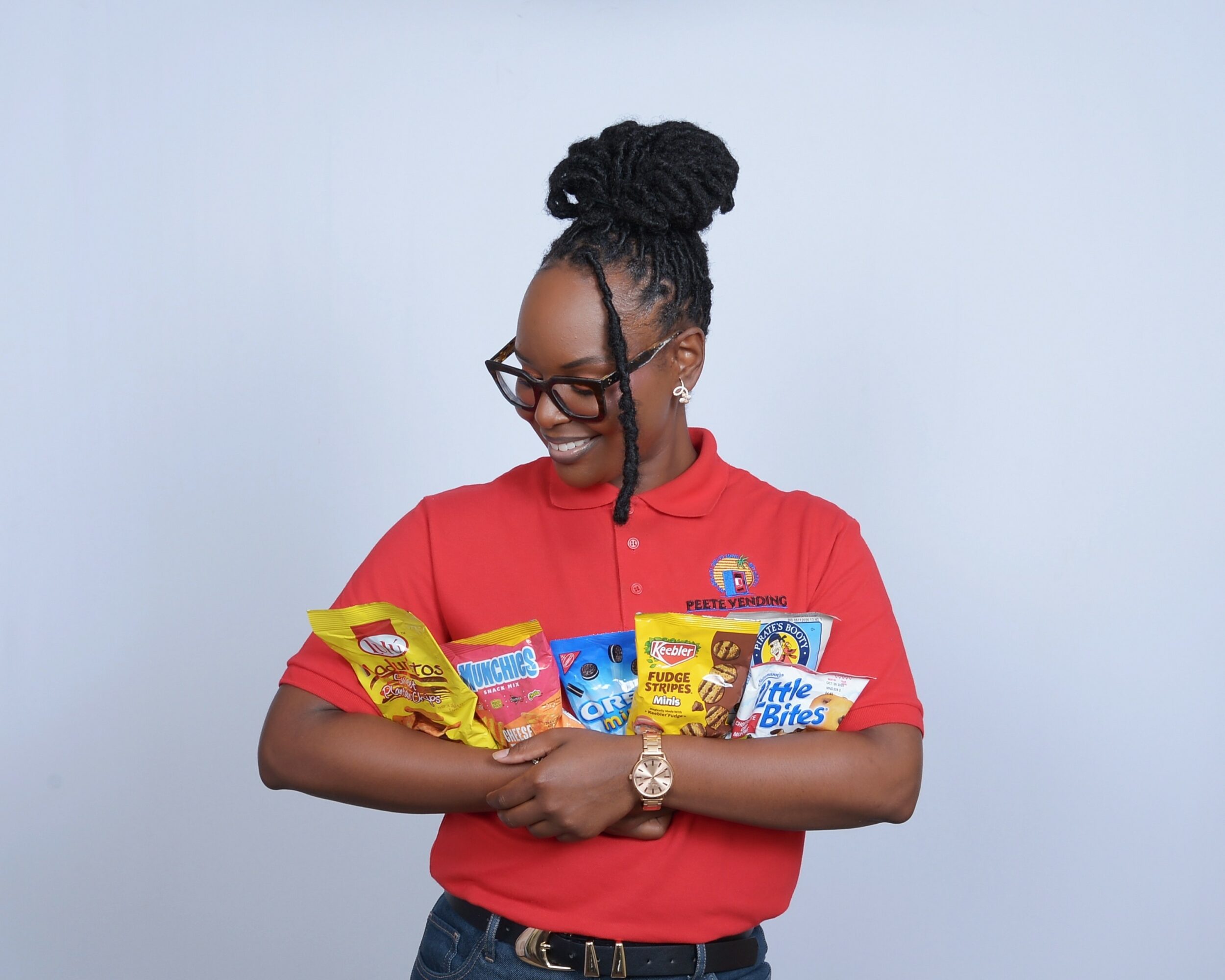 Woman with glasses and braided hair in a bun, smiling, holding snack bags, wearing a red shirt, against a light background.