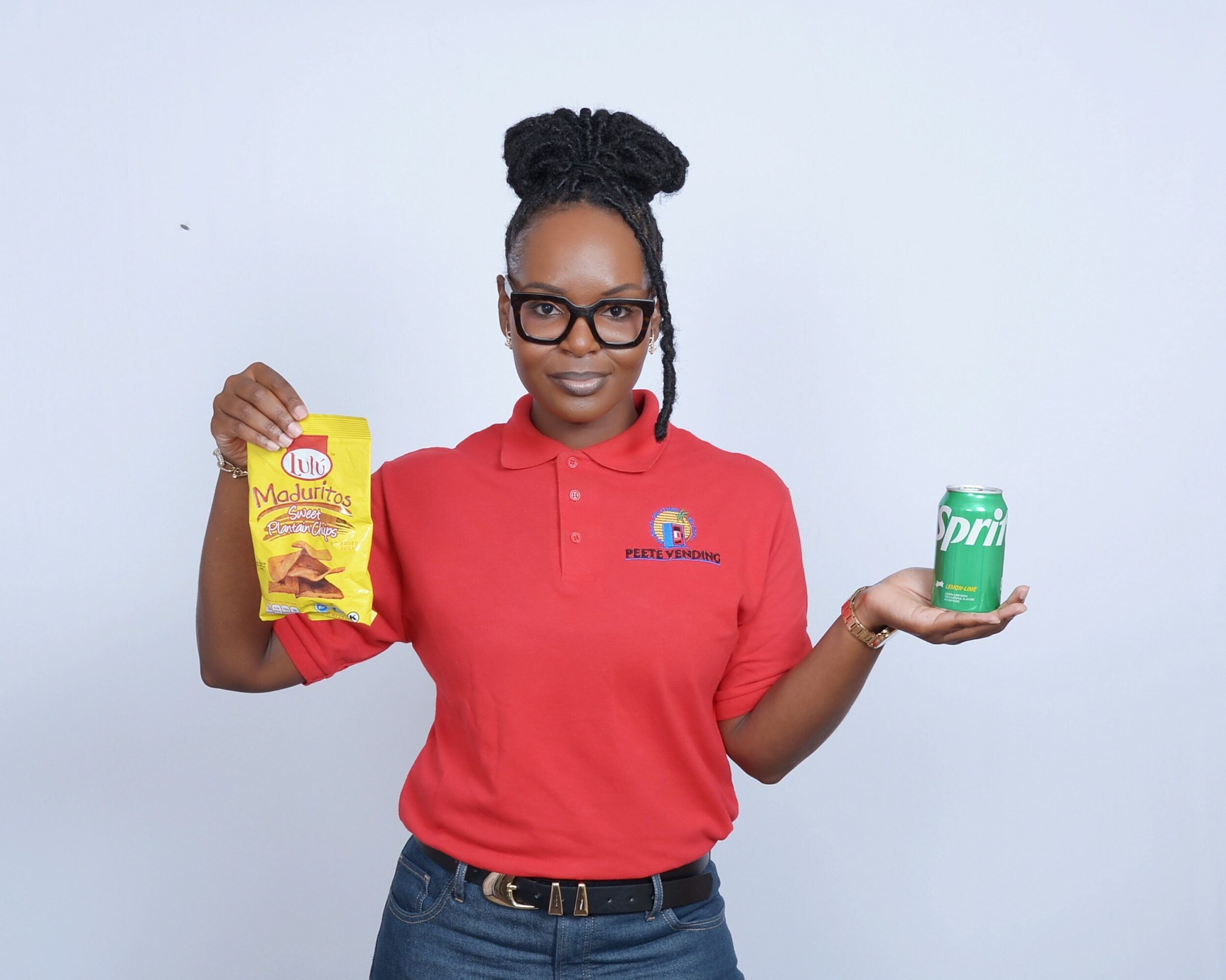 Woman with glasses in red polo shirt holding snack bag in one hand and soda can in the other, smiling.