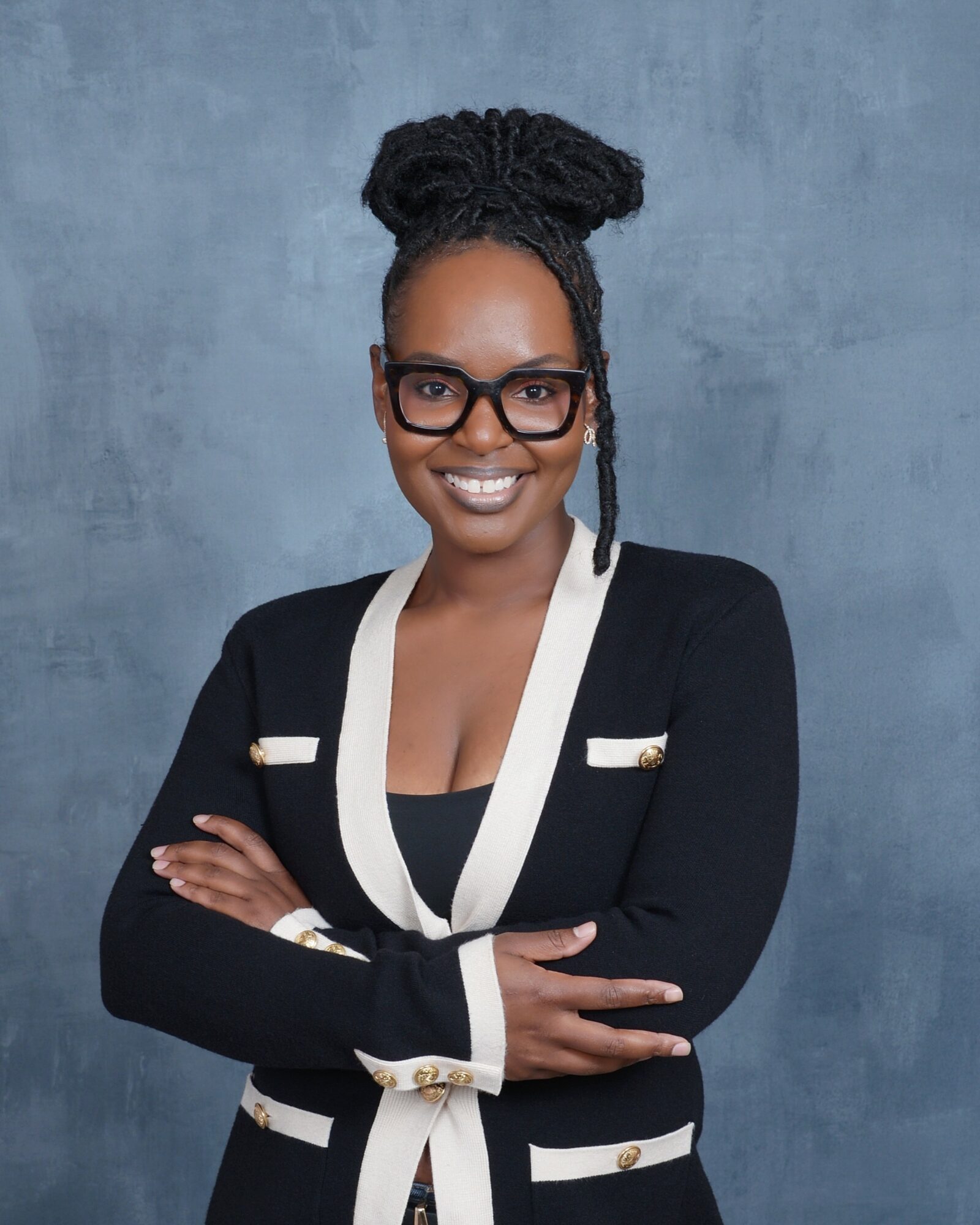 Woman with glasses and braided hair smiling, arms crossed, wearing a black and white blazer, standing against a blue background.