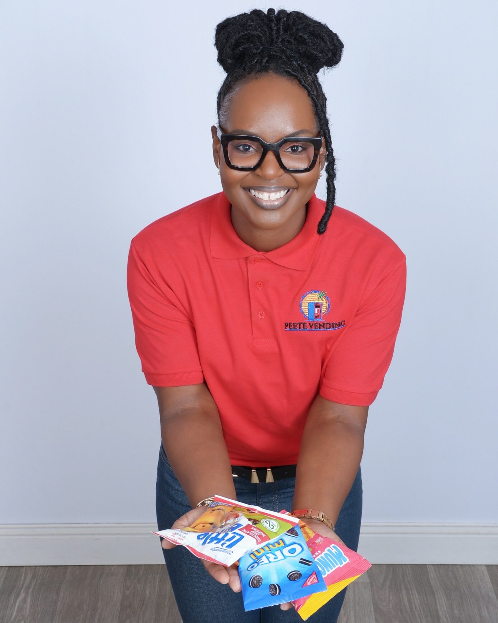 Smiling woman with glasses and braided hair, wearing a red polo shirt, holding a colorful magazine, standing against a plain background.