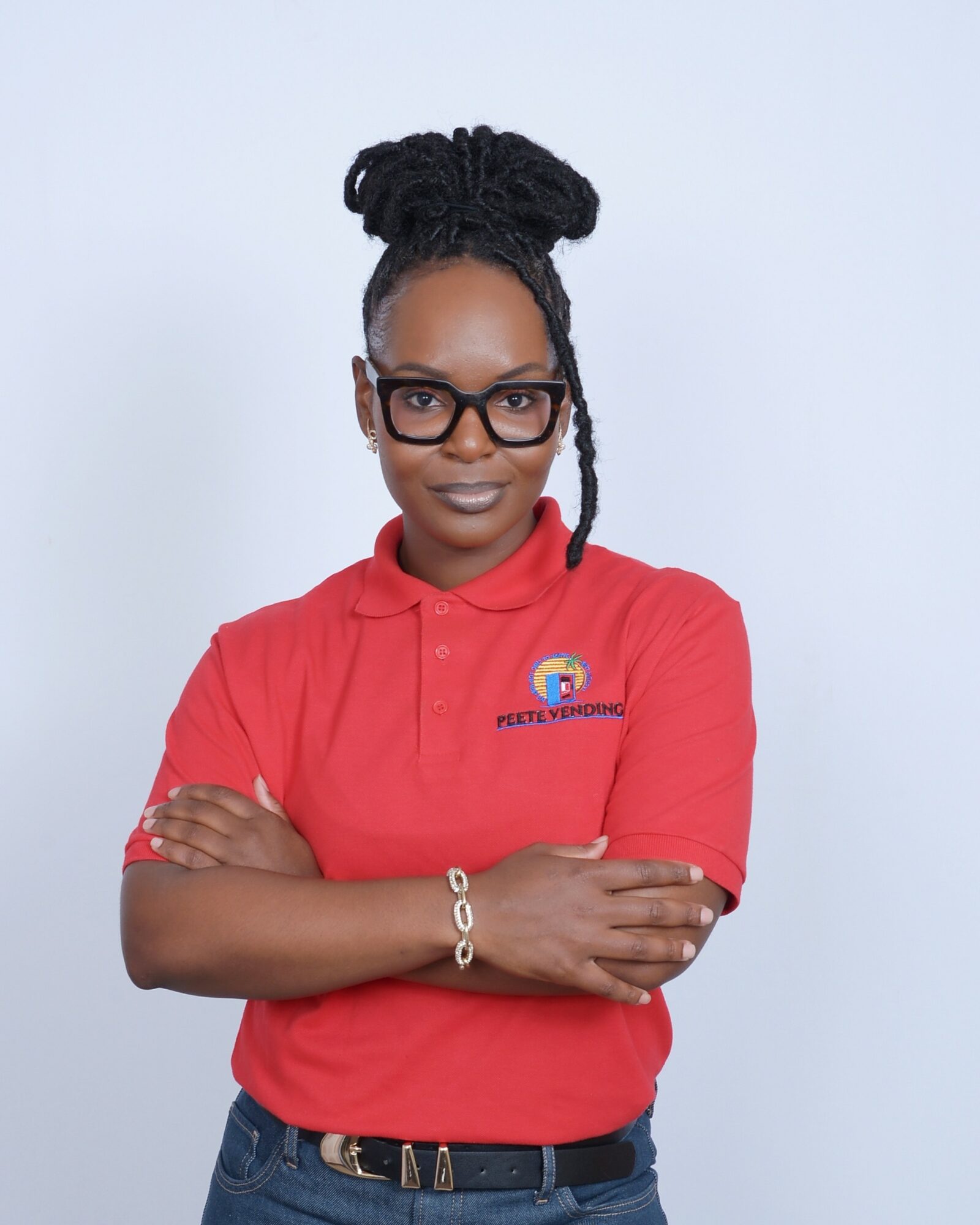 Woman with glasses and braided hair wearing a red polo shirt with a logo, arms crossed, smiling, against a light background.