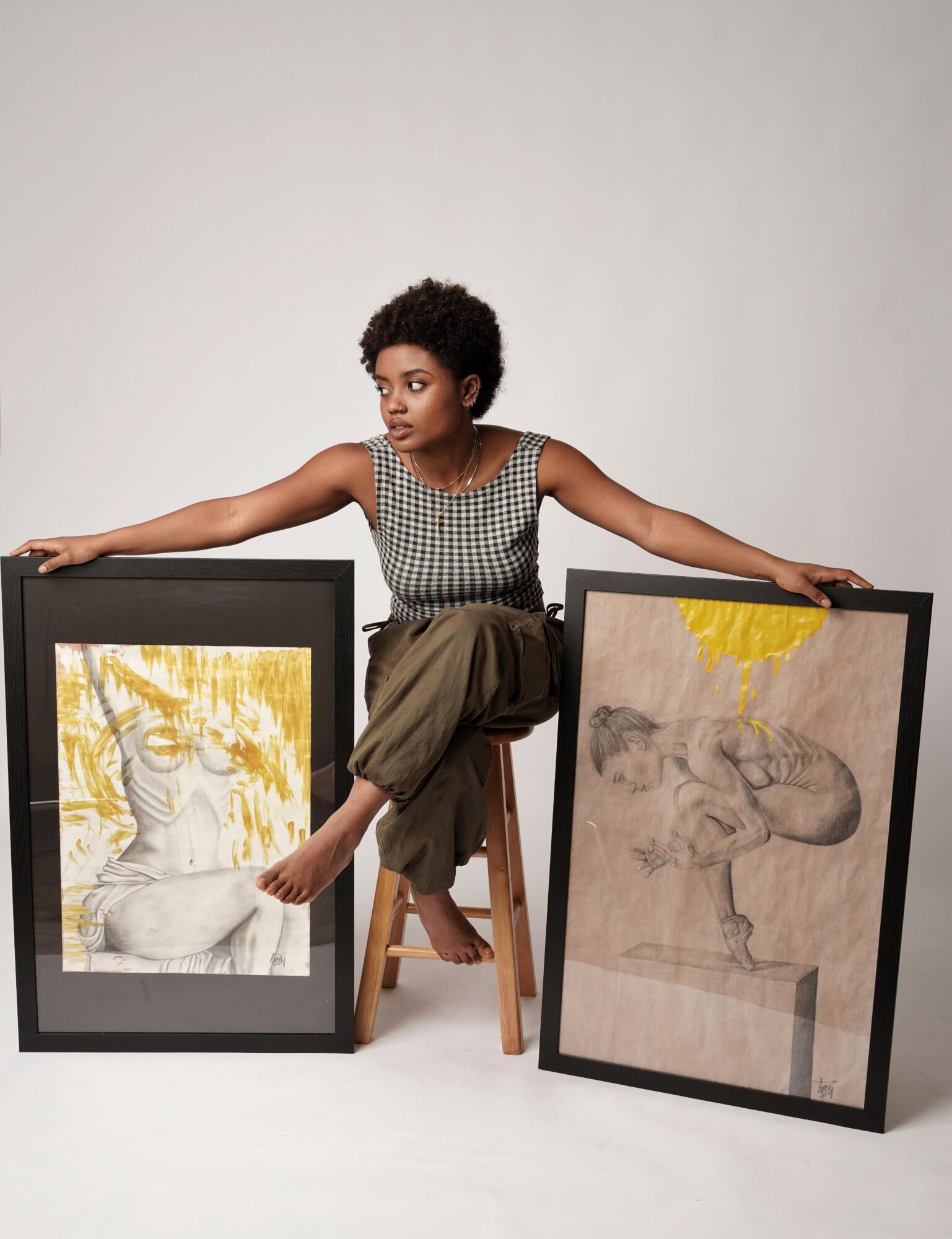 Young woman with short curly hair sitting on a stool, holding two framed artworks, one of a lion and one of a dog, against a plain background.