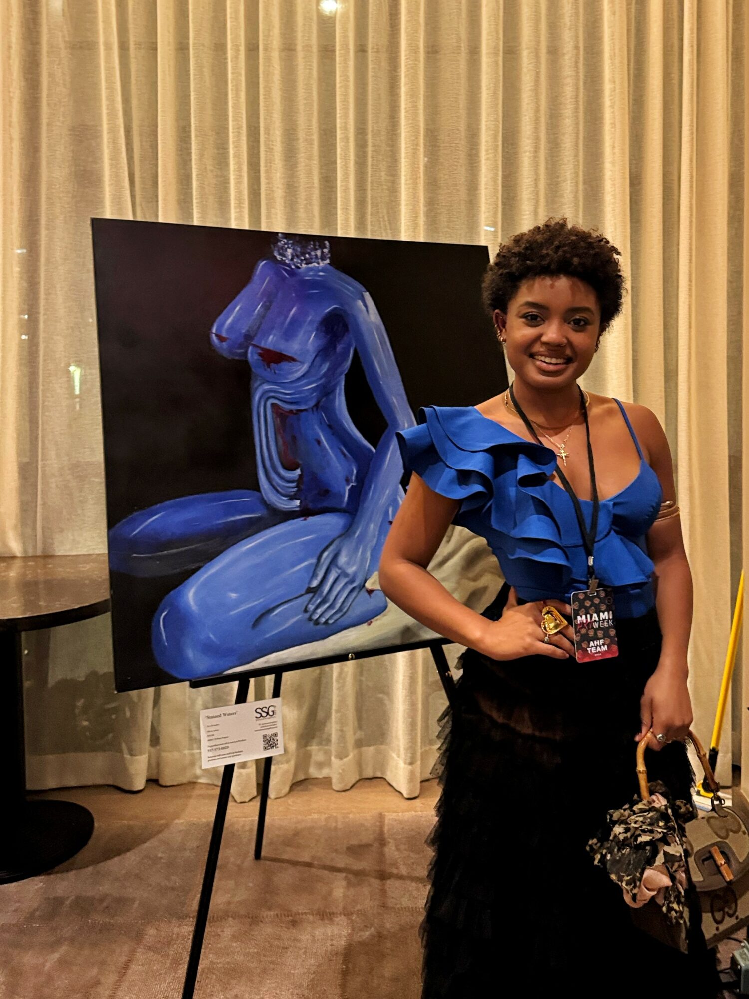 Woman with short curly hair smiling, wearing a blue ruffled top and black skirt, standing next to a blue sculpture on display.