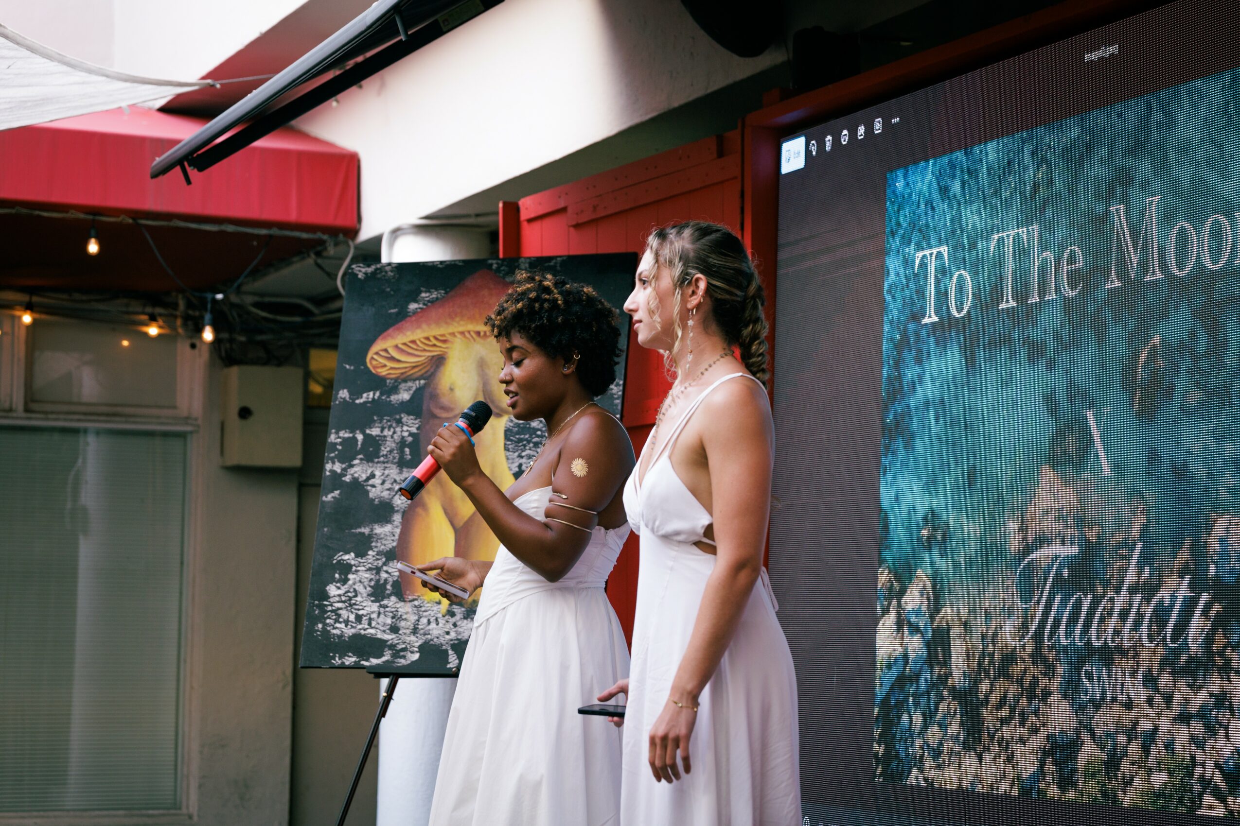 Two women in white dresses stand on stage, one speaking into a microphone, with a large screen and poster behind them.