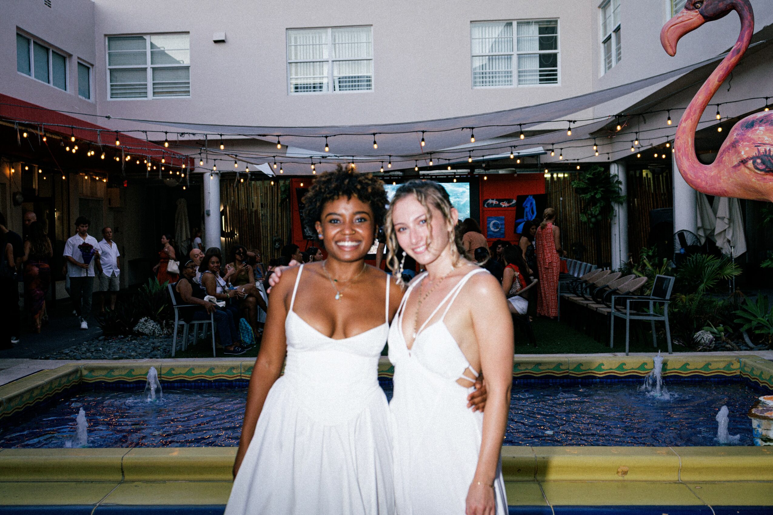 Two women in white dresses smiling outdoors near a fountain, with a pink flamingo statue and a building in the background.