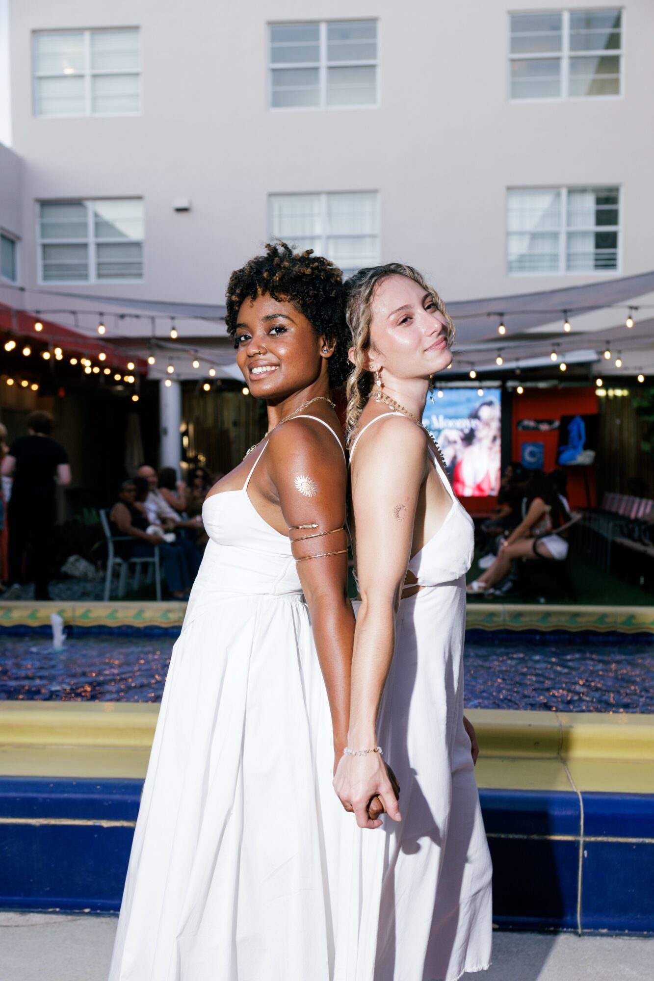 Two women in white dresses stand back-to-back outdoors at night, smiling, with a building and string lights in the background.