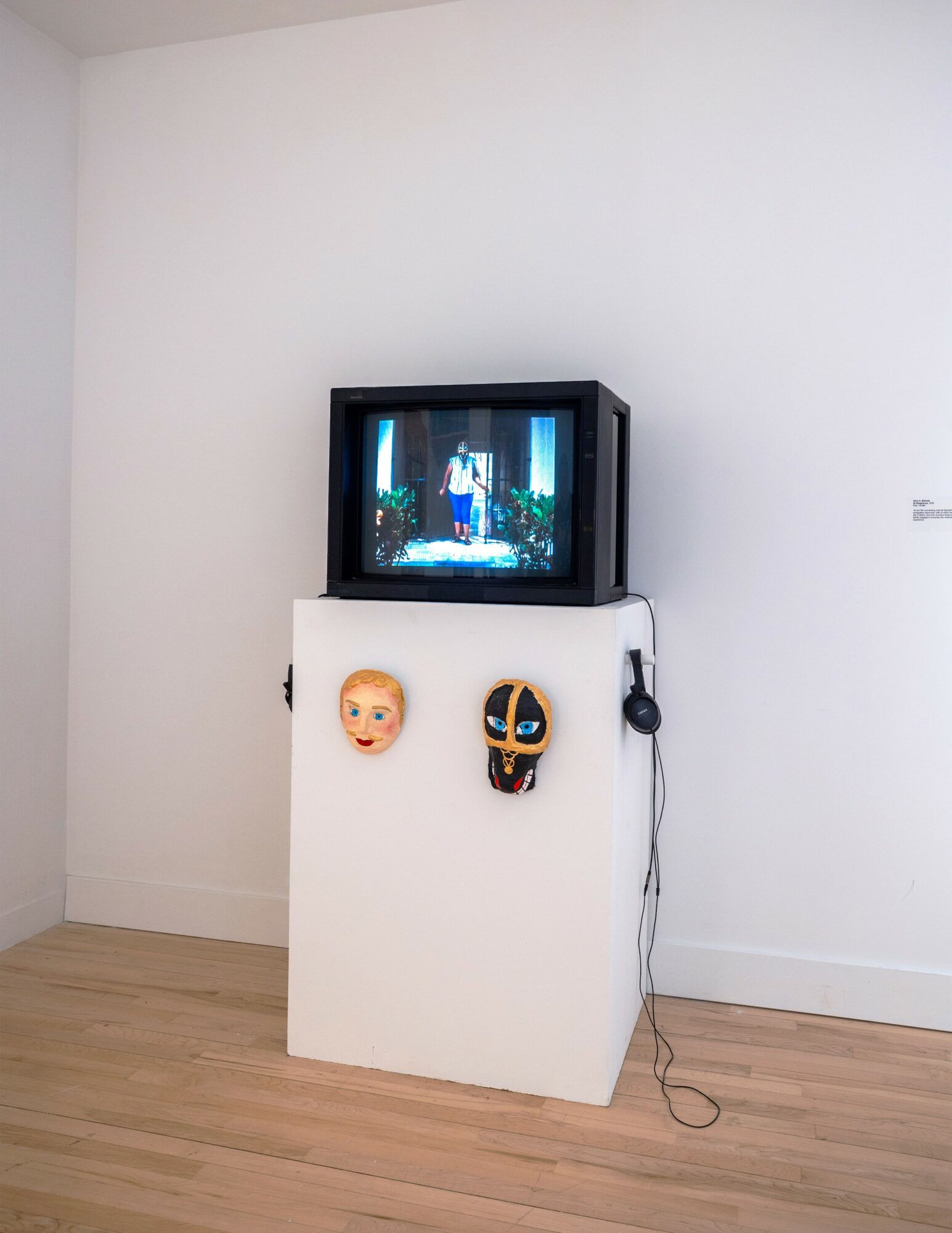 Television on a white pedestal displaying a scene, with a mask and a doll's face on the pedestal, and a headphone hanging.