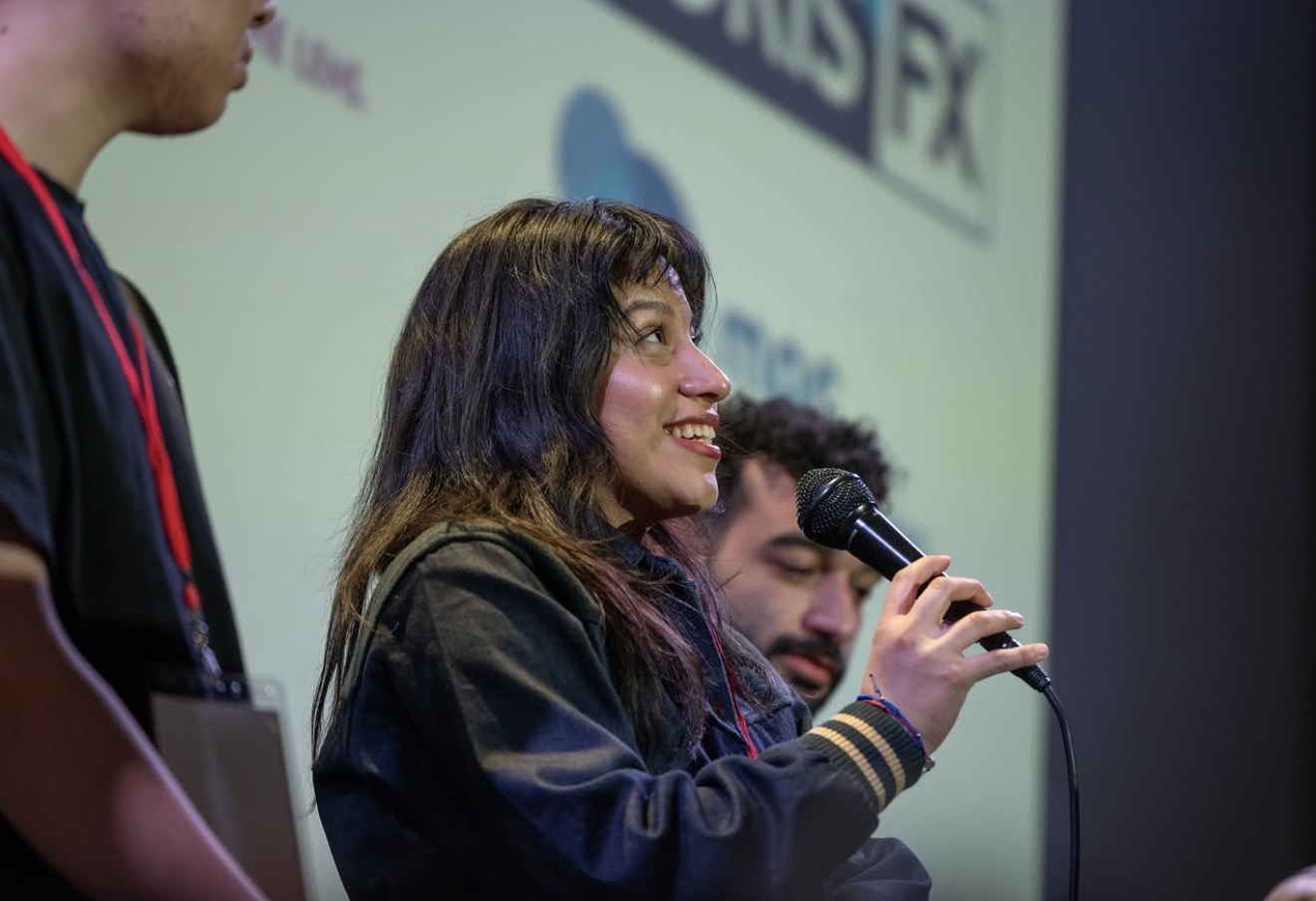 Woman speaking into a microphone during a panel discussion, with a man beside her and a presentation screen in the background.